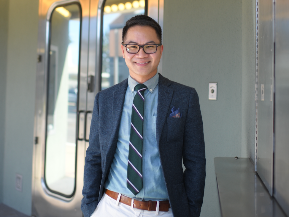 A smiling man with glasses, dressed in a business casual outfit, standing inside a building near a door and locker.