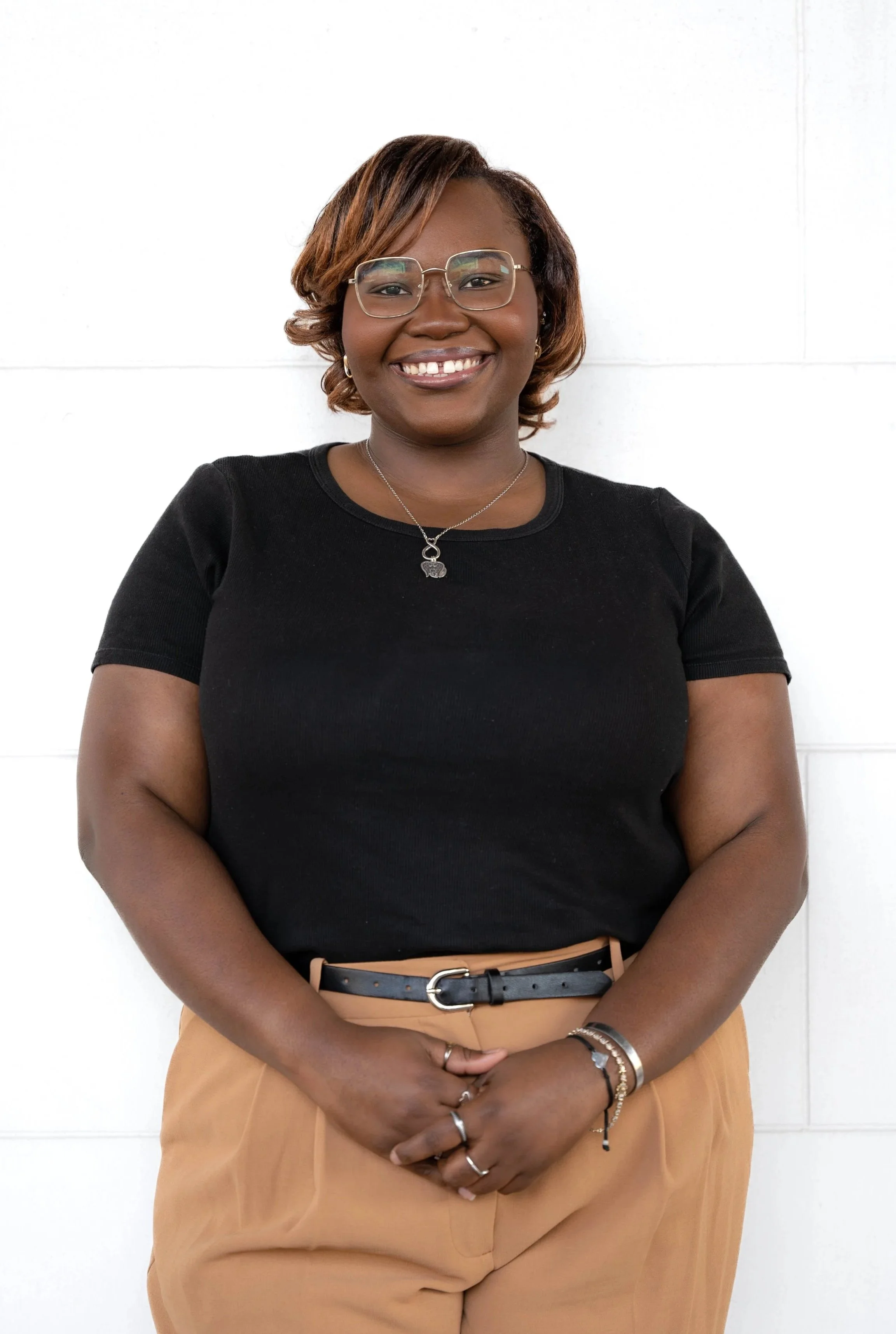 A woman with short hair, glasses, and dark skin, smiling and standing against a white wall. She is wearing a black t-shirt, beige pants, and several jewelry pieces including rings, bracelets, a necklace, and earrings.