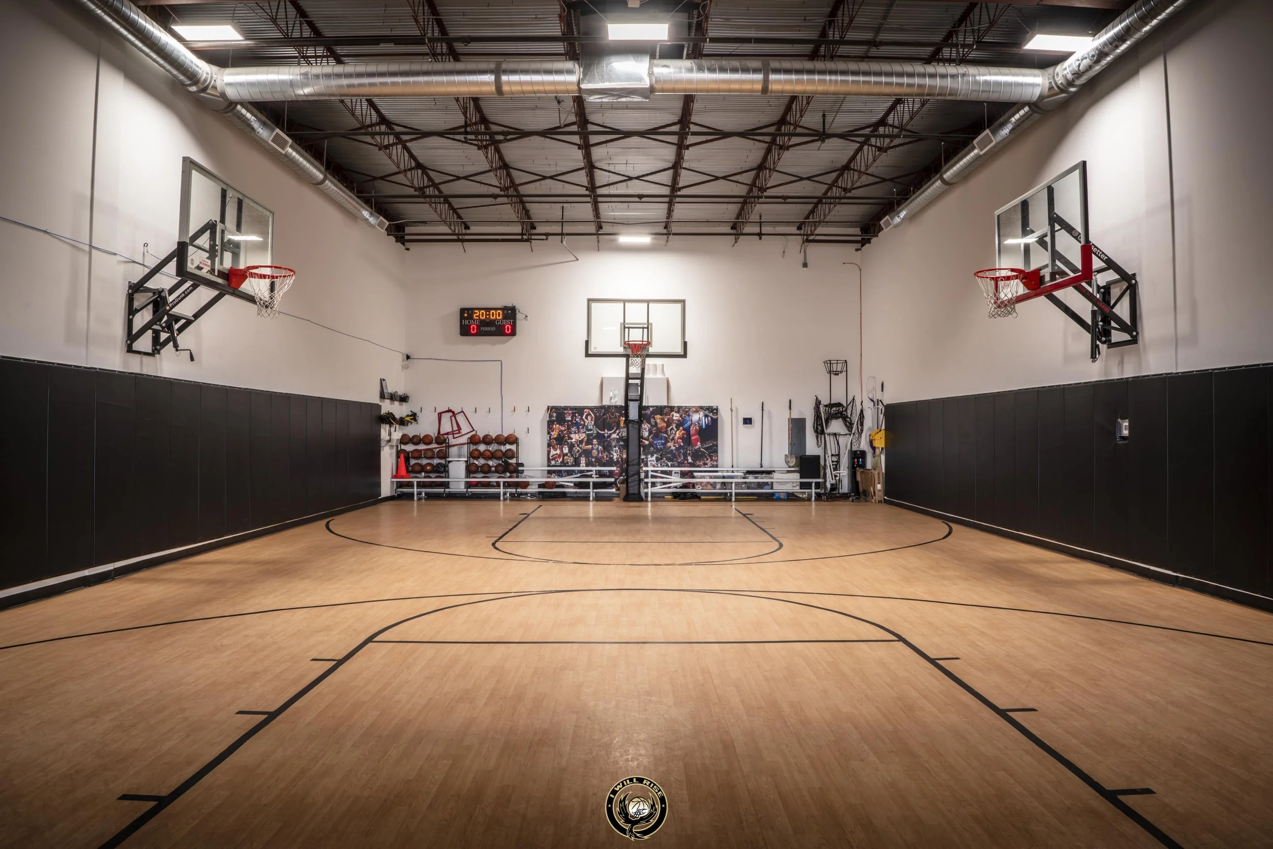Empty indoor basketball court with four basketball hoops, a scoreboard displaying 20:00, and gym equipment on the sidelines.