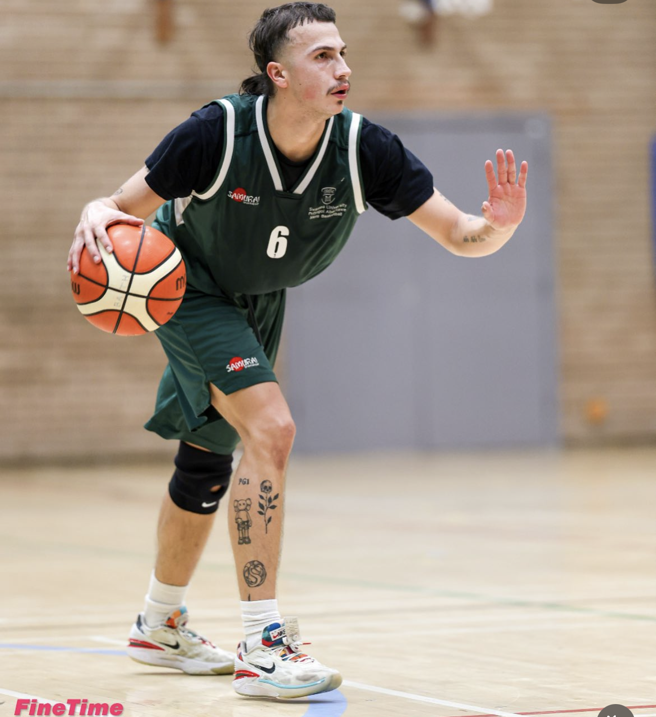 Young man with short, tousled brown hair, standing against a gray textured wall, wearing a black T-shirt with a logo and the words 'Moneyball Tour' and a gold chain necklace.