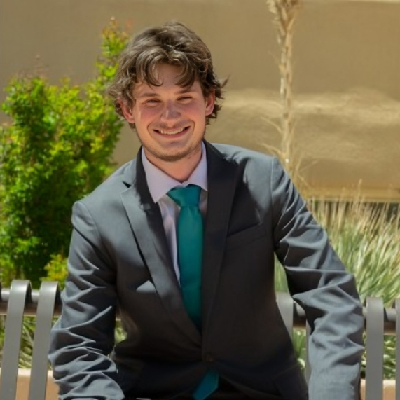 Young man in a gray suit and teal tie sitting on a bench outdoors, smiling at the camera.