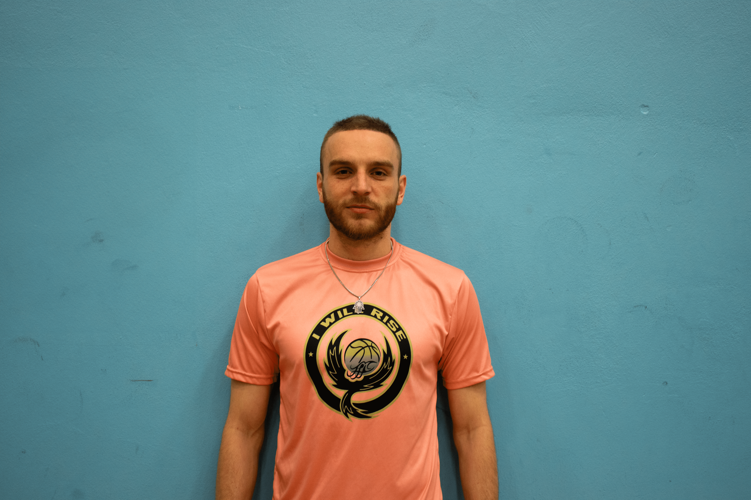 A young man with short dark hair and a beard wearing a pink t-shirt with a black and gold logo and a silver necklace, standing against a turquoise wall.