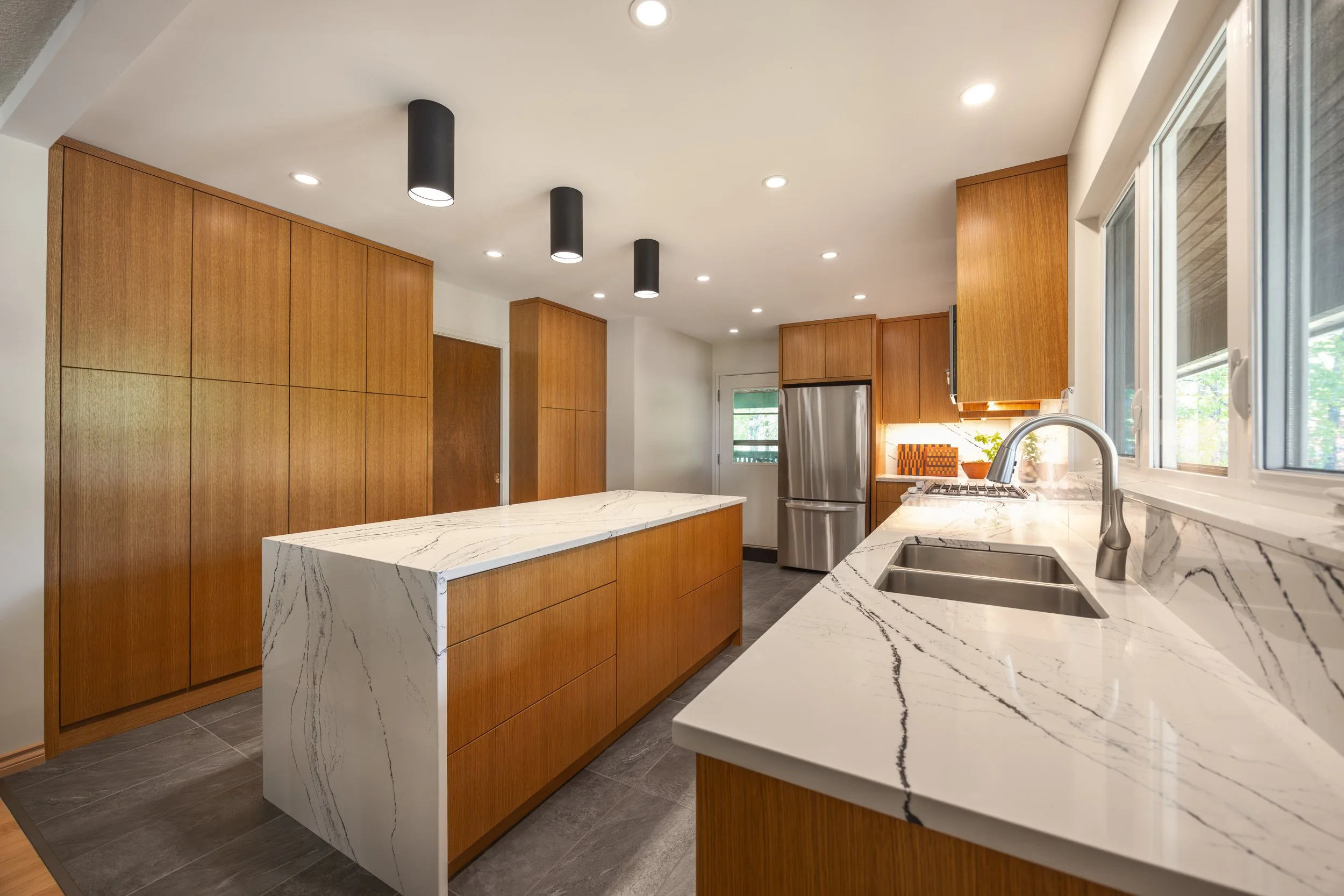 Modern kitchen with wooden white oak cabinets, marbled quartz countertops, stainless steel refrigerator, and black surface mount lights.
