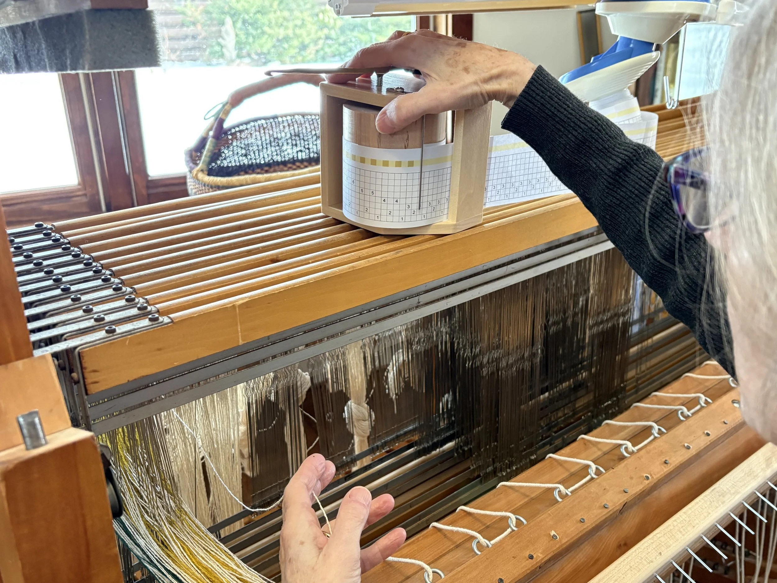 Weaver's hand moving a cylinder that has graph paper, depicting a threading draft for weaving