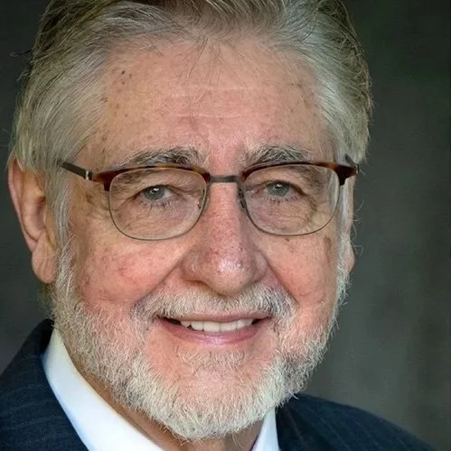 Close-up portrait of Dr. Craighead with gray hair and beard, wearing glasses and a dark suit, smiling at the camera.