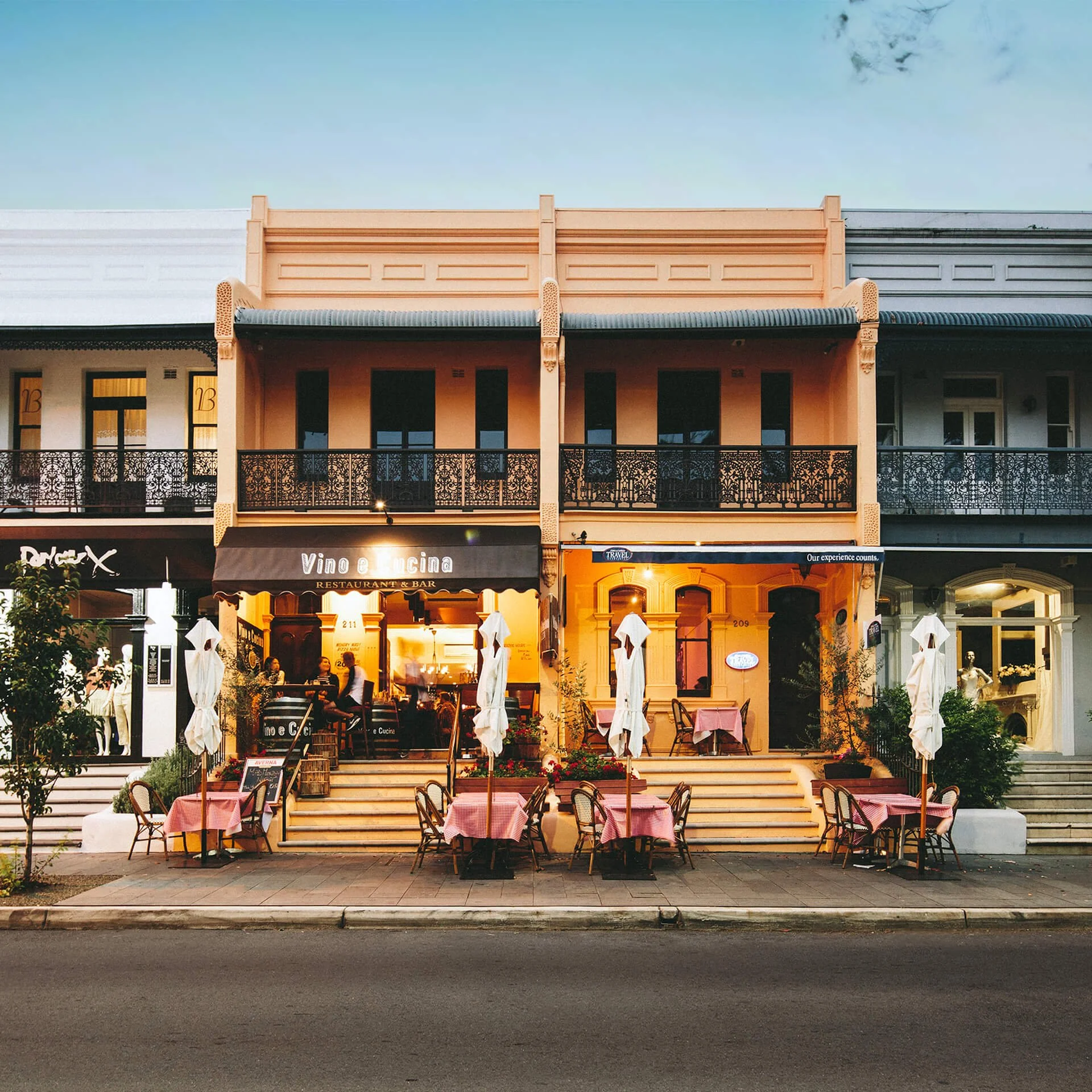 Front view of a two-story restaurant with outdoor seating on stairs and sidewalk, pink tablecloths, umbrellas, and warm lighting, with neighboring storefronts on either side.