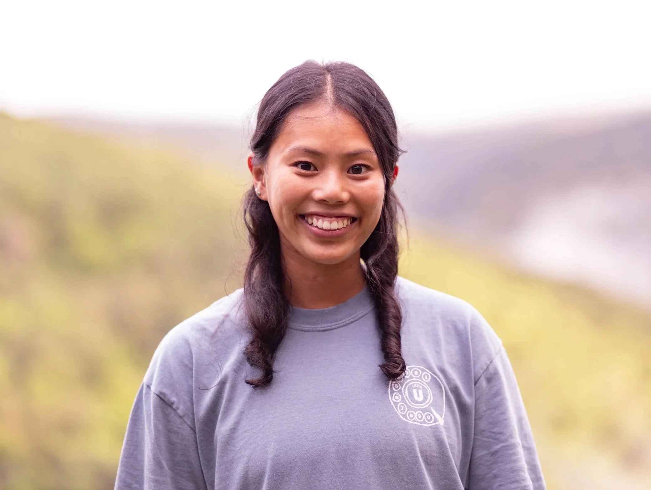 A young woman with long braided hair, wearing a gray sweatshirt with a circular logo on the chest, outdoors with a blurred natural background.