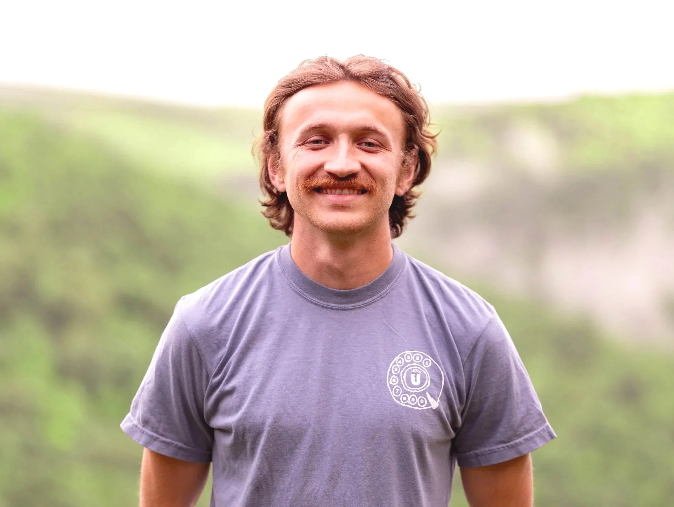 A young man with light skin, curly light brown hair, and a mustache, smiling outdoors. He is wearing a gray T-shirt with a small circular logo on the left side that reads 'Listen U,' against a background of greenery and a cloudy sky.