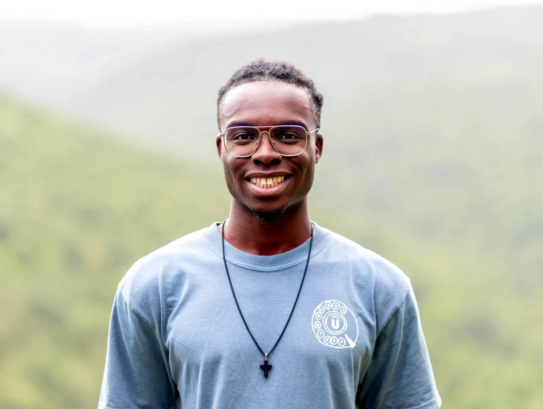 Smiling young man with glasses, wearing a blue T-shirt and a necklace, standing outdoors on a foggy day.