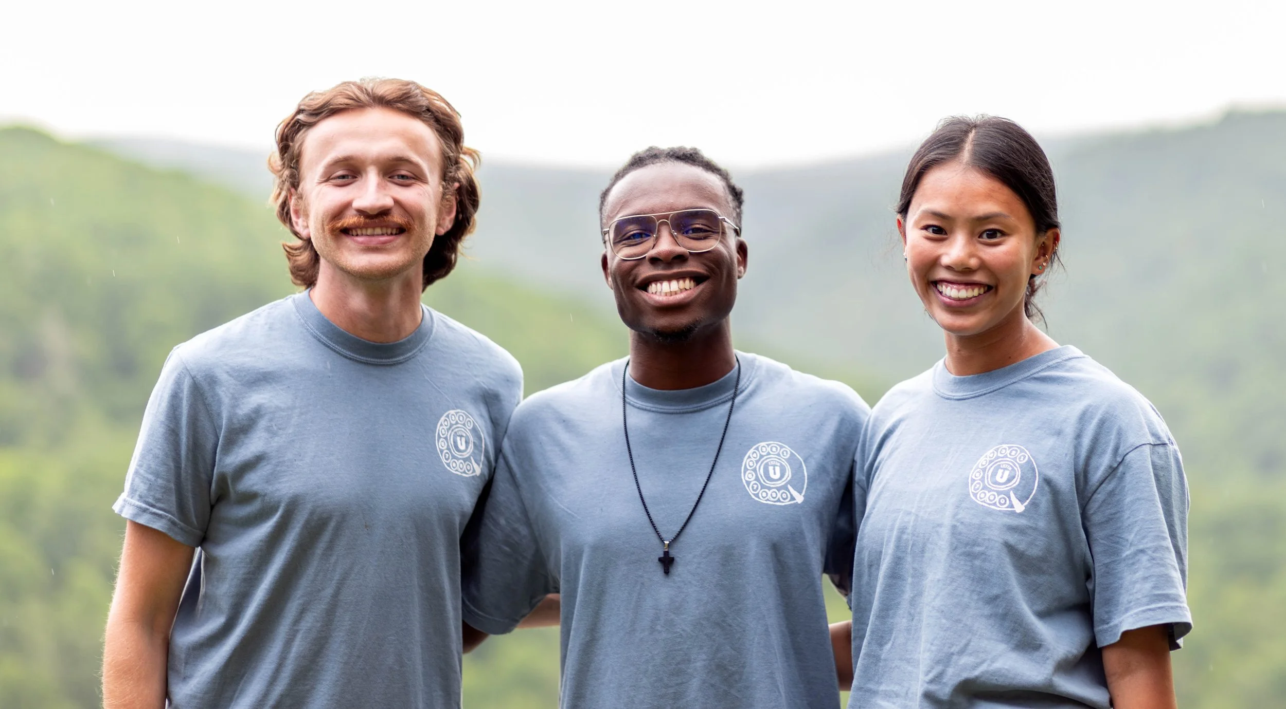 Three young adults standing outdoors in casual T-shirts, smiling at the camera with a blurred natural background.