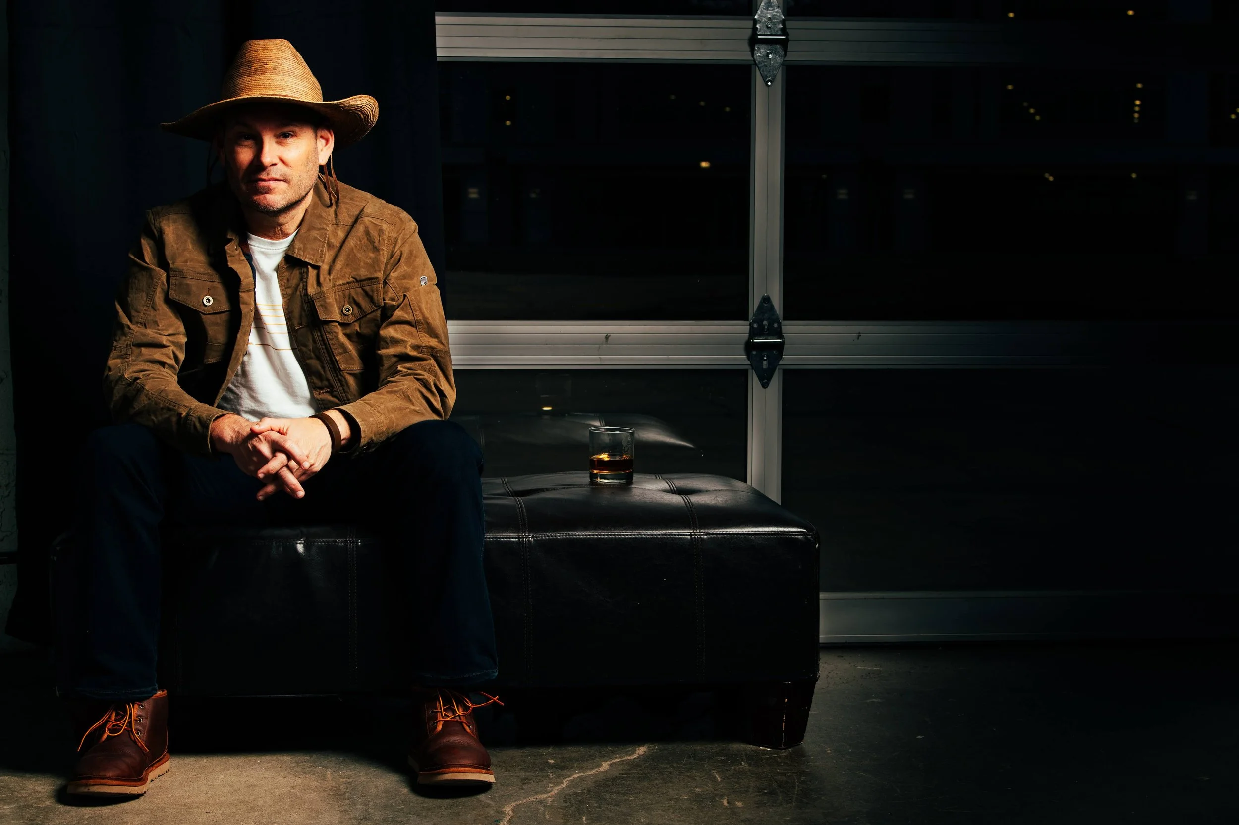 A man sitting on a black leather ottoman in a dimly lit room at night, wearing a wide-brimmed straw hat, brown jacket, white t-shirt, dark pants, and brown boots, with a glass of whiskey beside him.