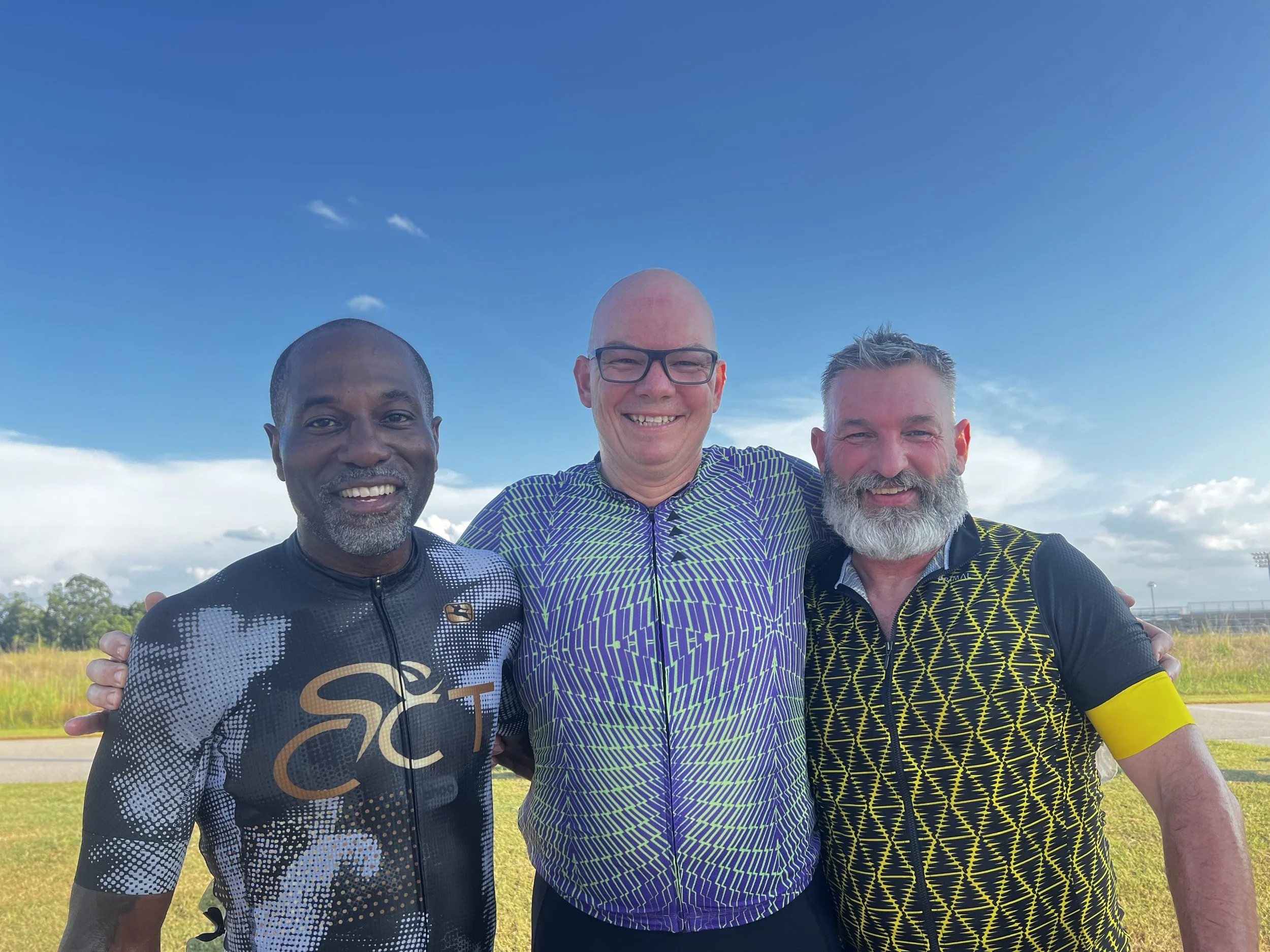 Three cyclists riding bikes on a paved road, viewed through banners on either side in a park or open area with green trees in the background.