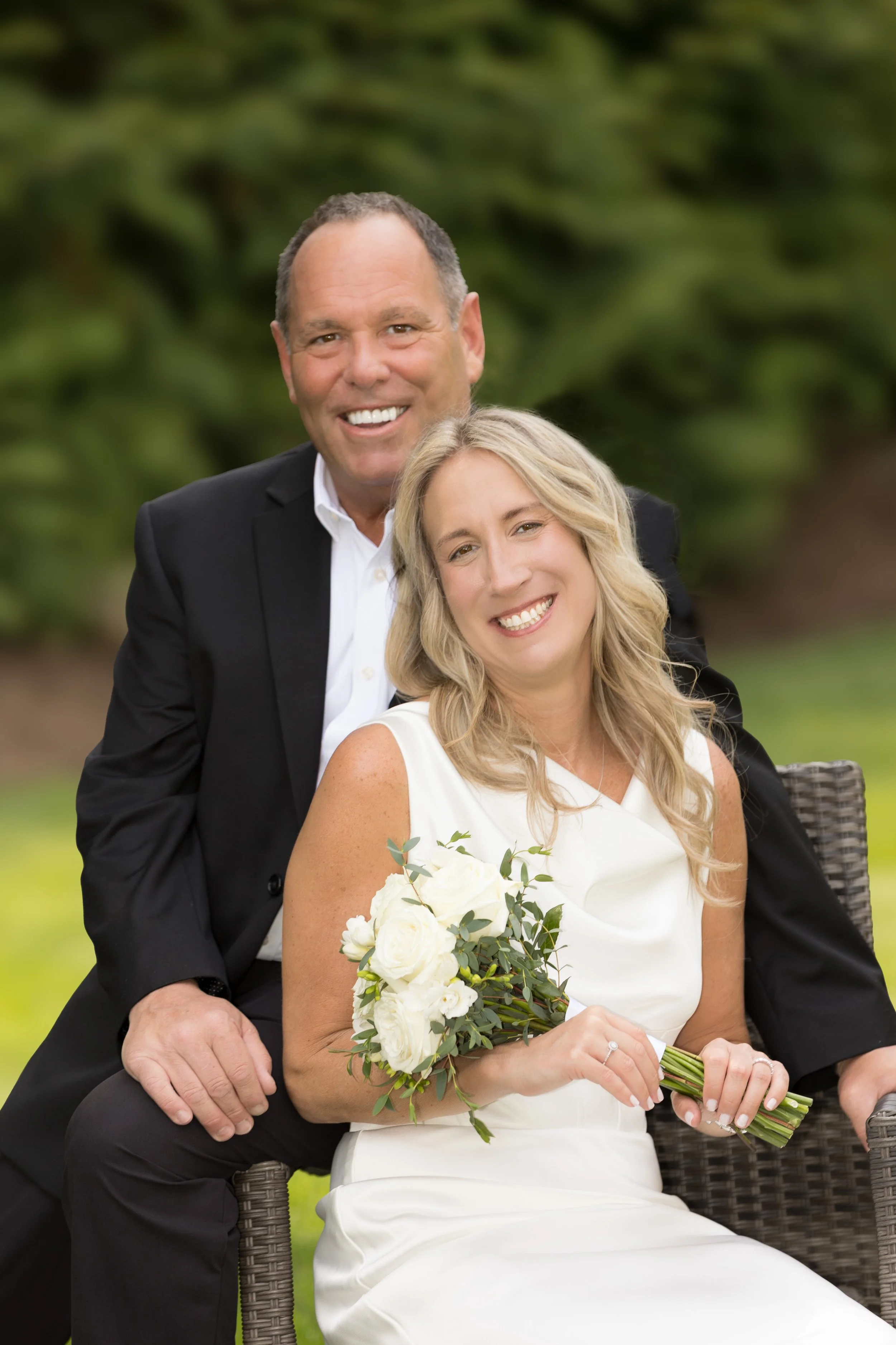 A smiling middle-aged man in a black suit and white shirt with a woman in a white dress sitting outdoors. The woman is holding a bouquet of white roses, and they are seated in a wicker chair with greenery in the background.