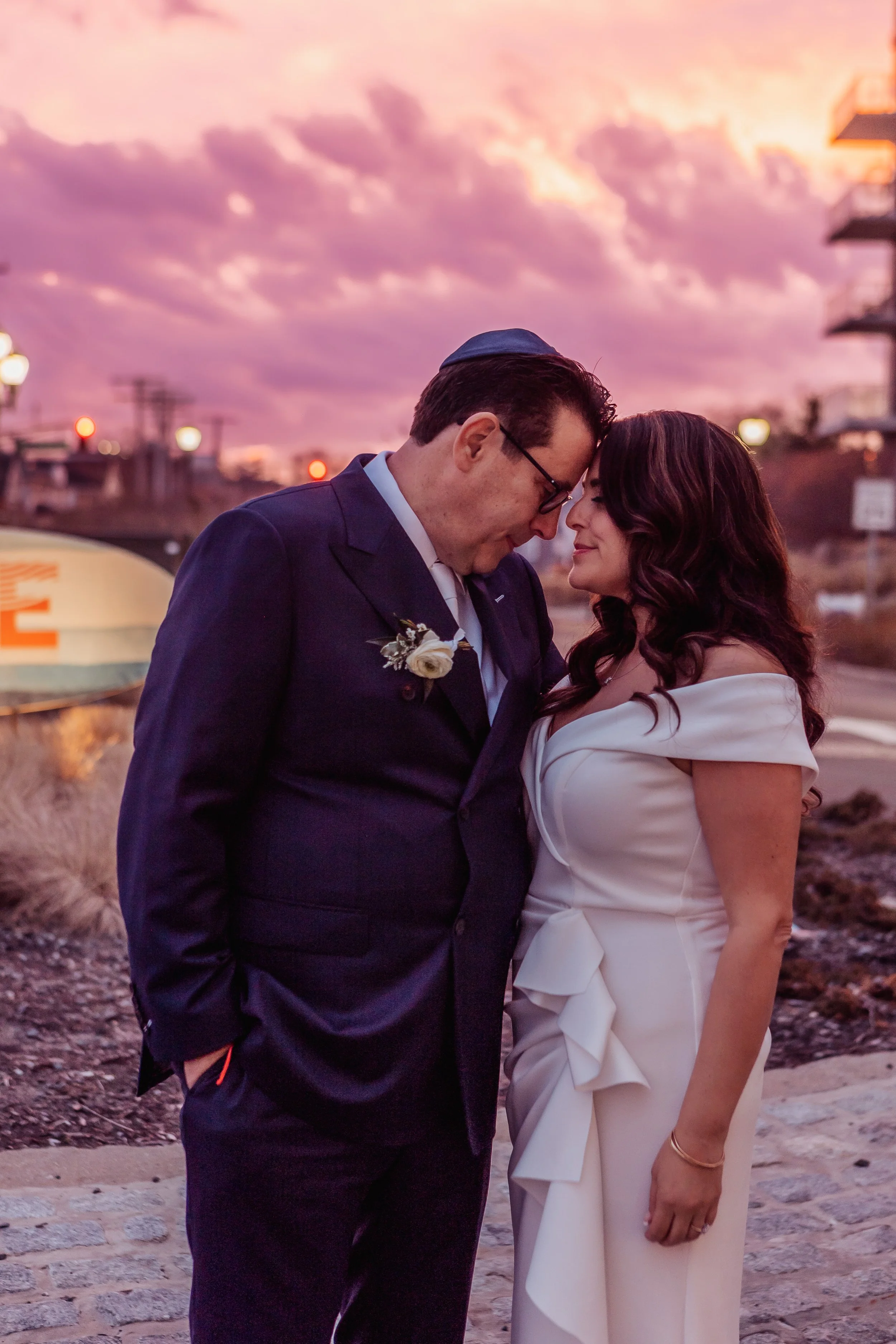 A couple in wedding attire sharing a tender moment at sunset, with their foreheads touching and eyes closed. The background features a colorful pink and purple sky with clouds and a blurred cityscape.