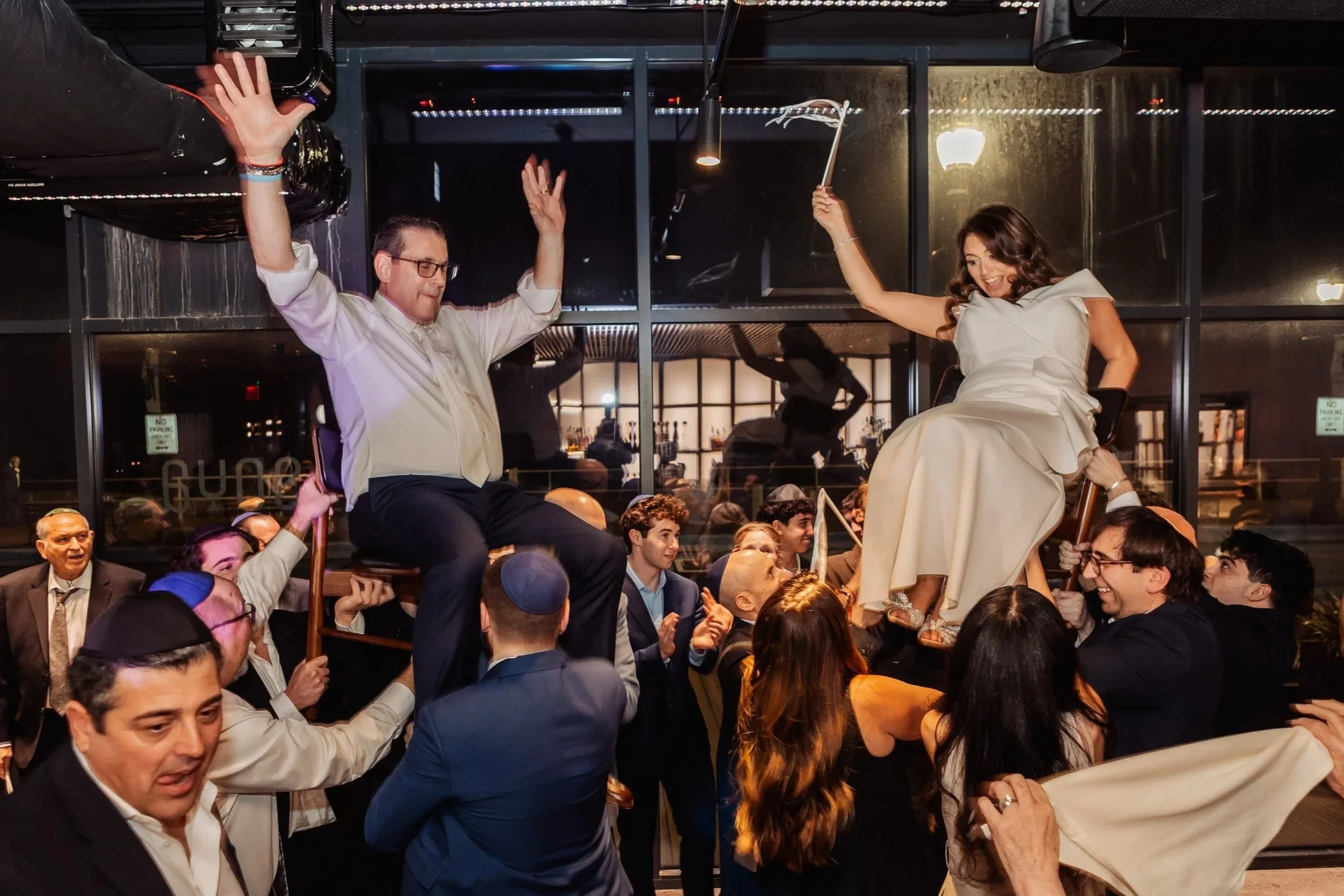 People are participating in a celebratory event, with two individuals on chairs elevated above the crowd. One of the individuals is a woman in a white dress, smiling and holding a long spoon, while the other is a man in a white shirt with glasses, ra