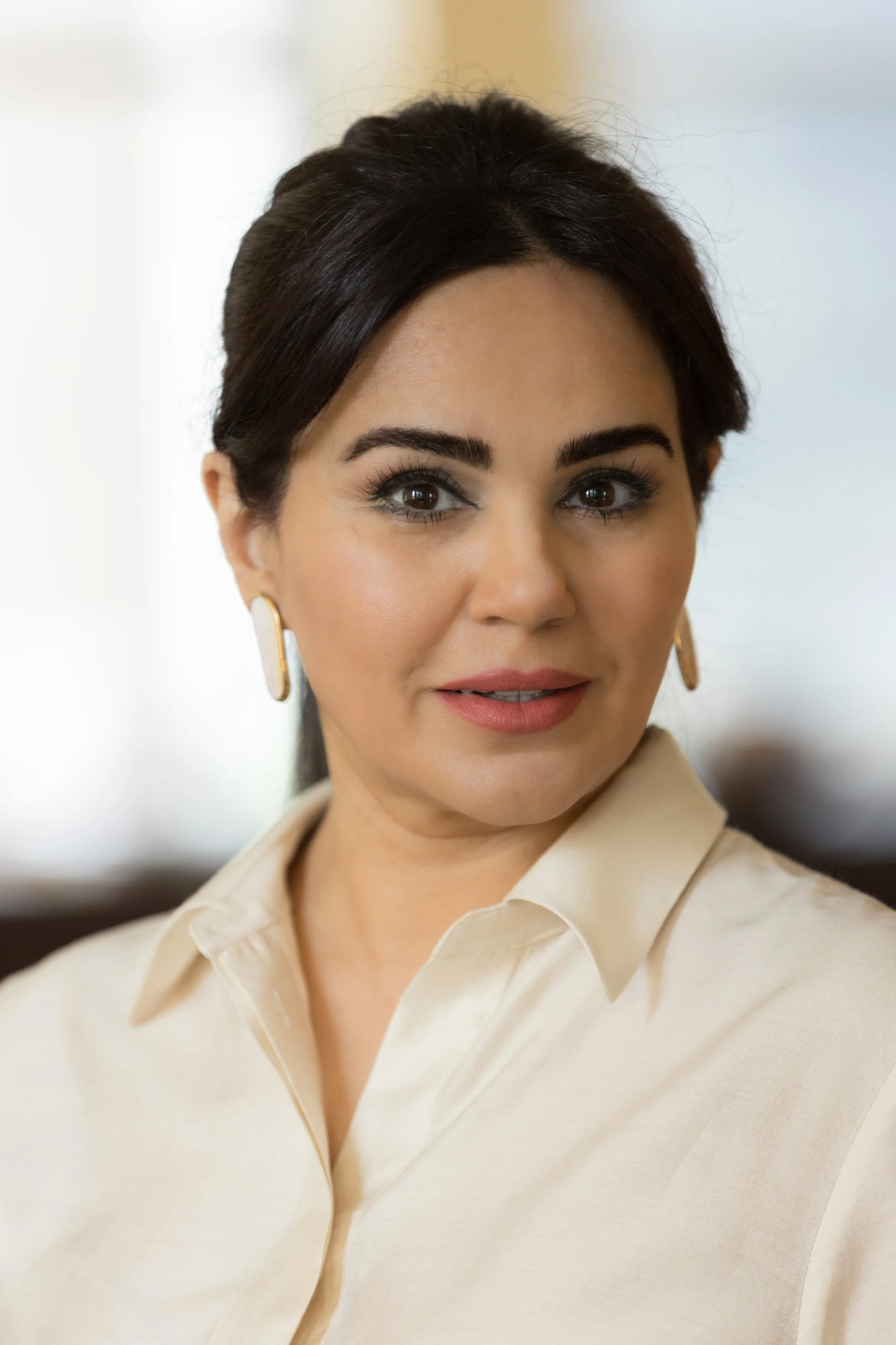 Close-up of a woman Maryam Shojai   with dark hair, wearing a cream-colored blouse and gold earrings, looking slightly to the side with a soft smile.