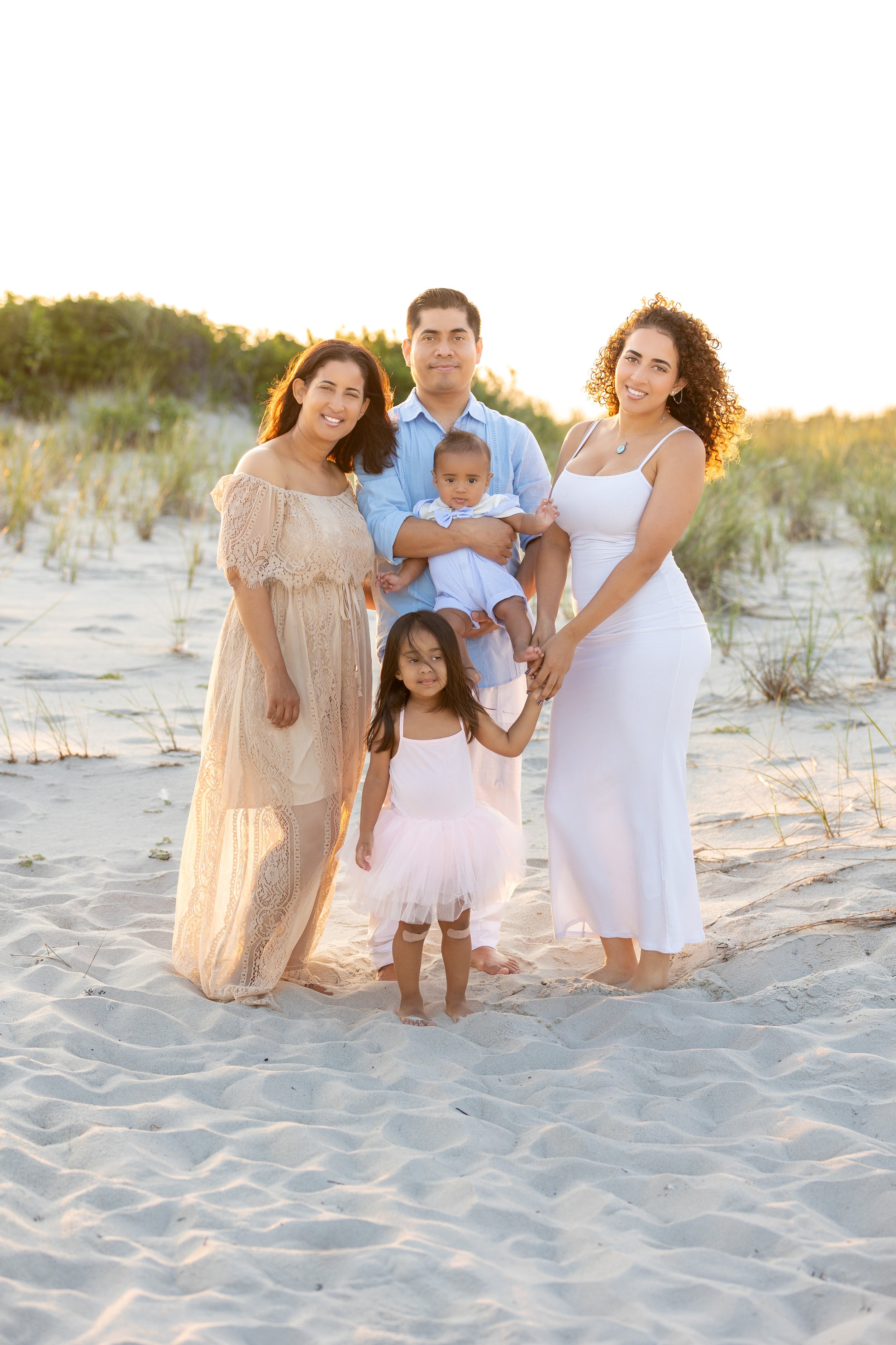 A multi-generational Hispanic family standing on a sandy beach during sunset, smiling at the camera. The group includes two women, a man holding a baby, and two young girls.