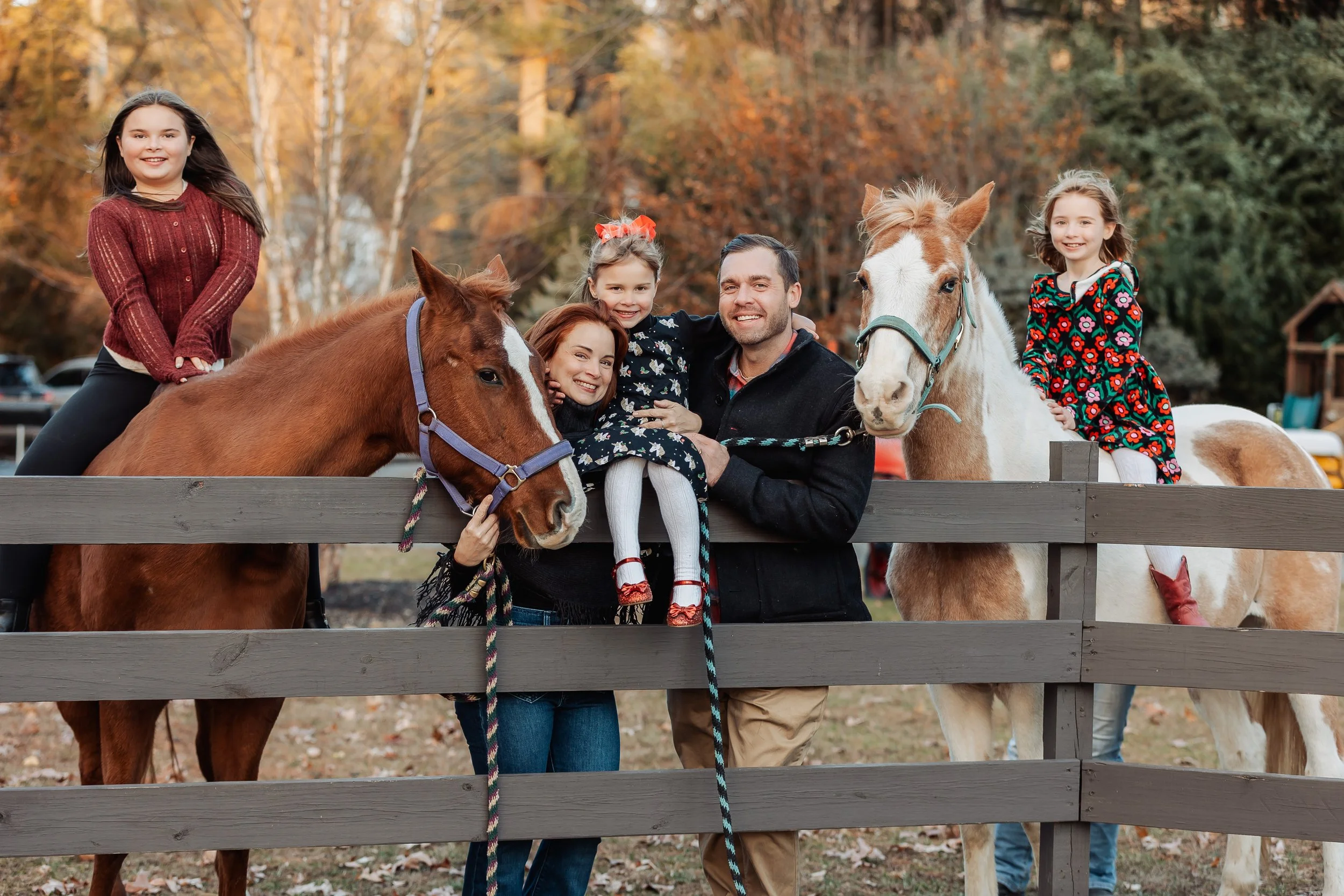 A group of people, including children and adults, standing behind a wooden fence with horses, in an outdoor setting with autumn trees in the background.