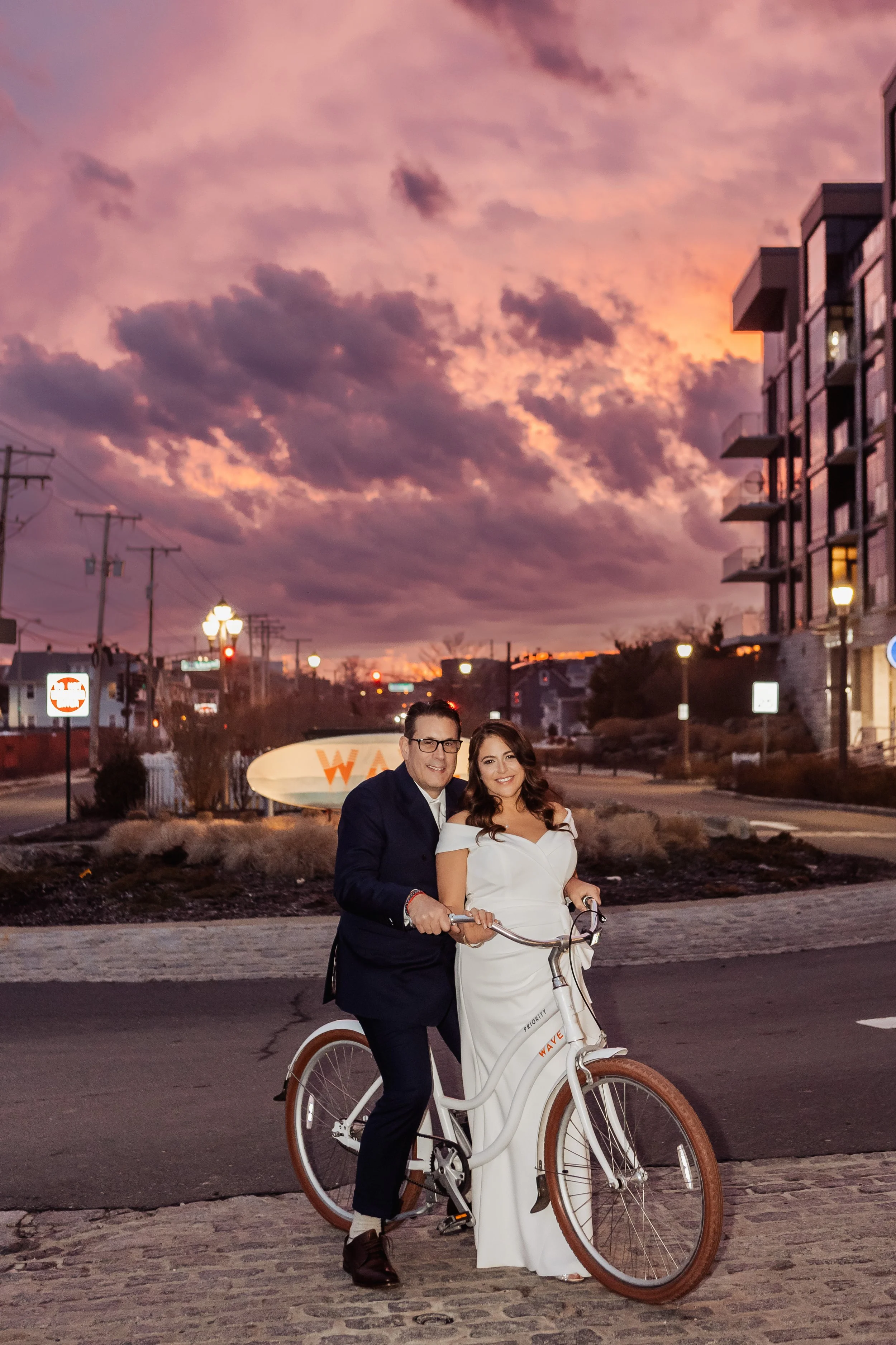 A couple dressed in formal attire, with the woman in a white gown and the man in a dark suit, riding a white pedal bicycle together on a cobblestone street at sunset. The sky is filled with pink and purple clouds, and there are buildings and streetli