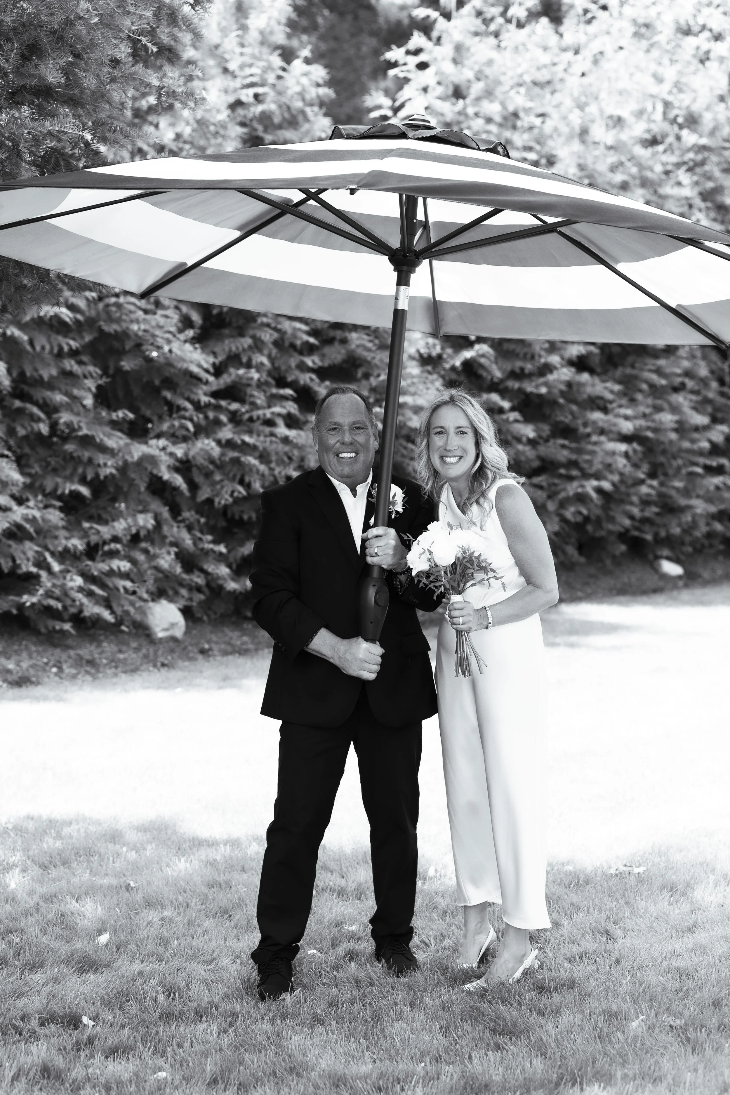 A man and a woman standing on grass beneath a large striped umbrella, smiling for the camera, with trees in the background.