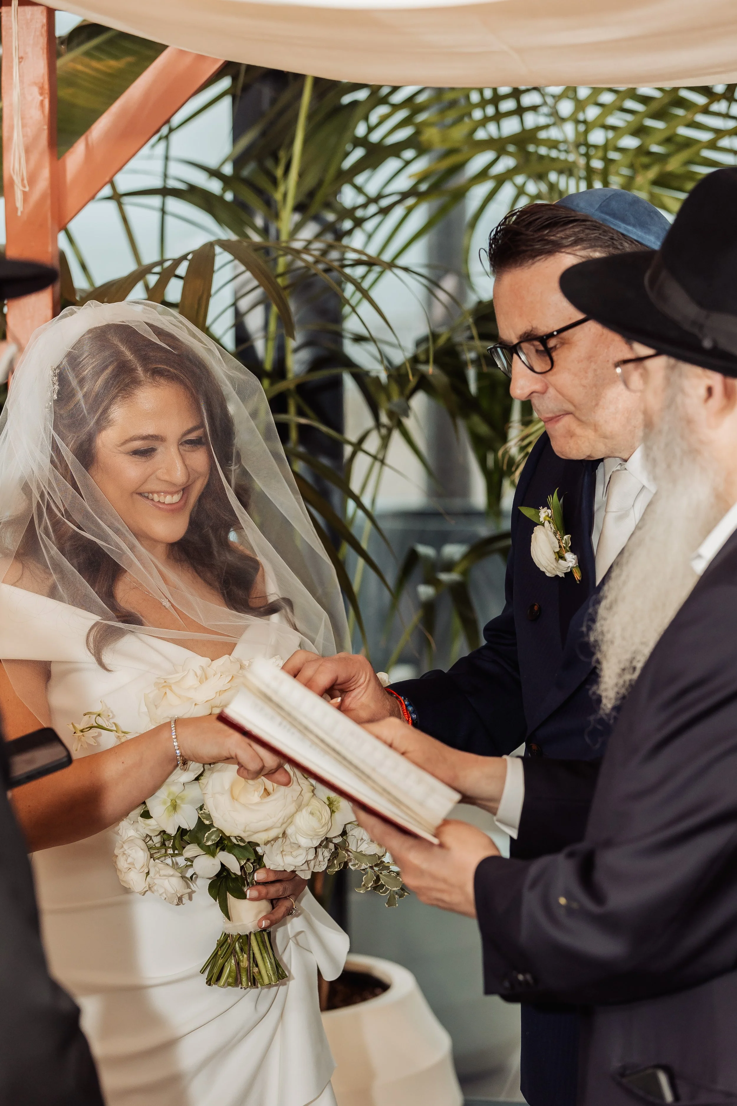 A bride with a veil and wedding dress holding a bouquet of white flowers, during her marriage vow exchange with a groom in a suit and tie, with a person in background reading from a book.