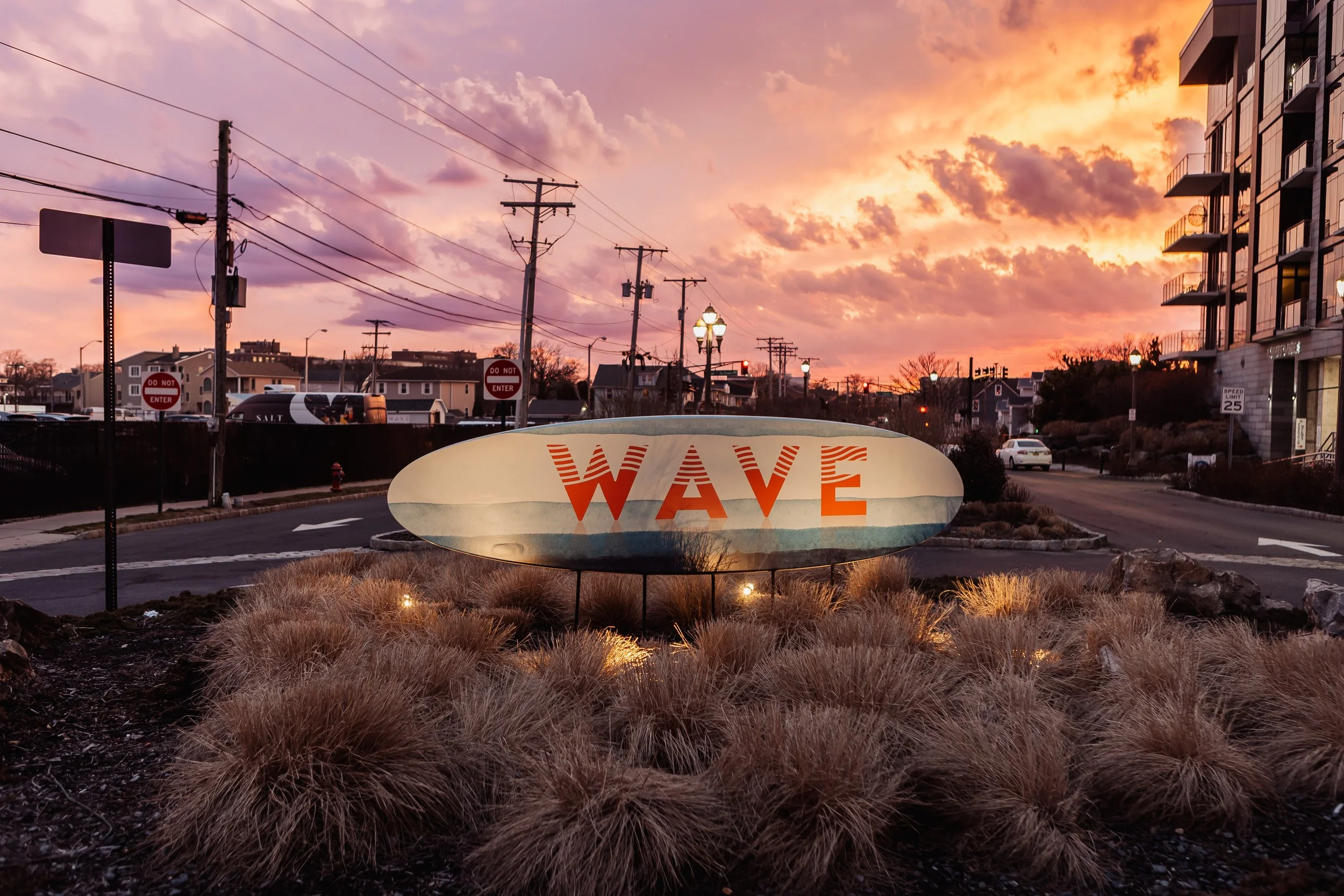 View of a sunset sky with pink and orange clouds over a street with utility poles, streetlights, and buildings, featuring a large decorative sign with the word 'WAVE' in orange.