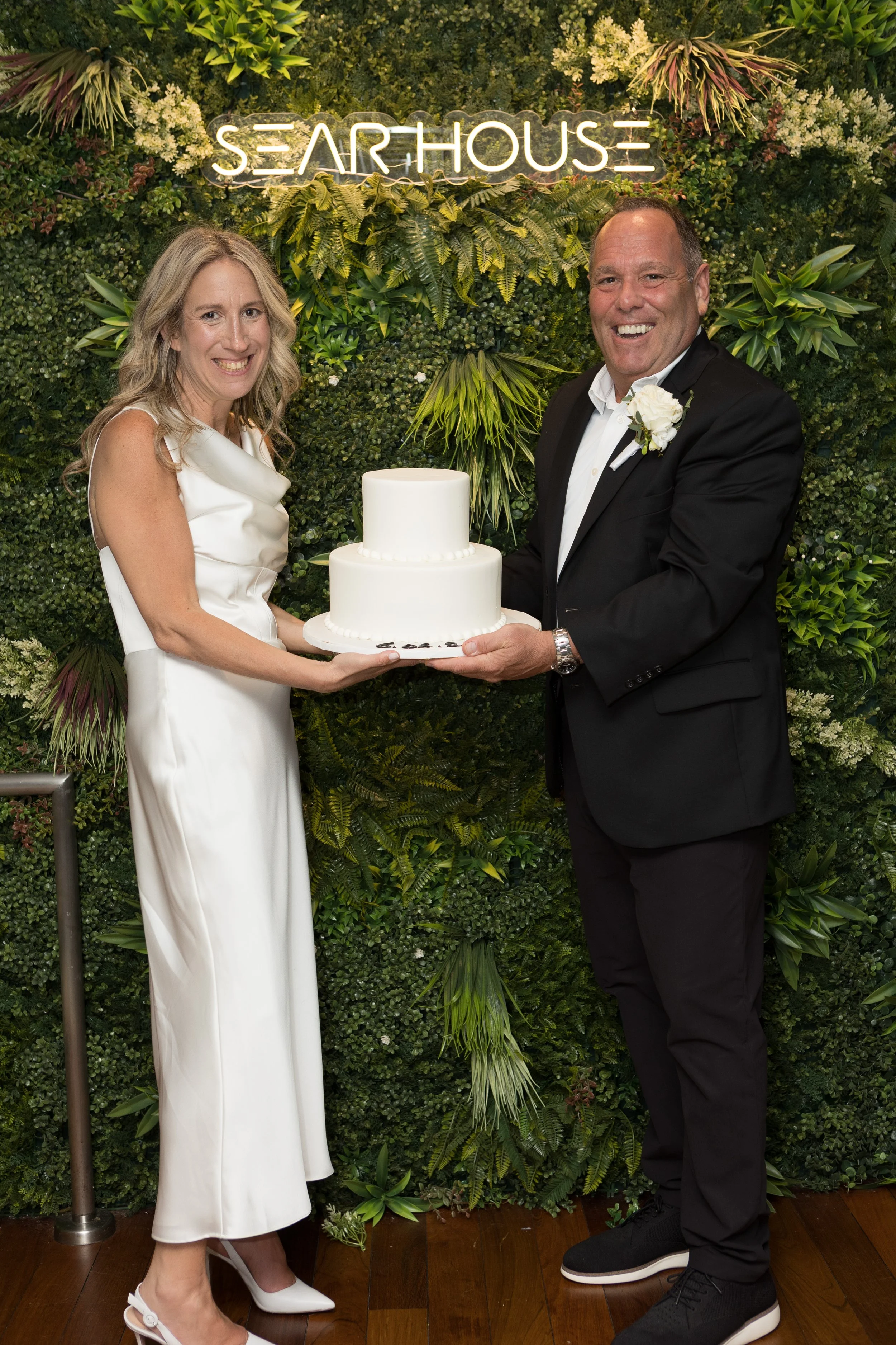 A bride and groom holding a wedding cake together at a wedding celebration. The bride is wearing a white dress and heels, and the groom is in a black suit with a white shirt and a white flower boutonniere. They are standing in front of a green leafy 