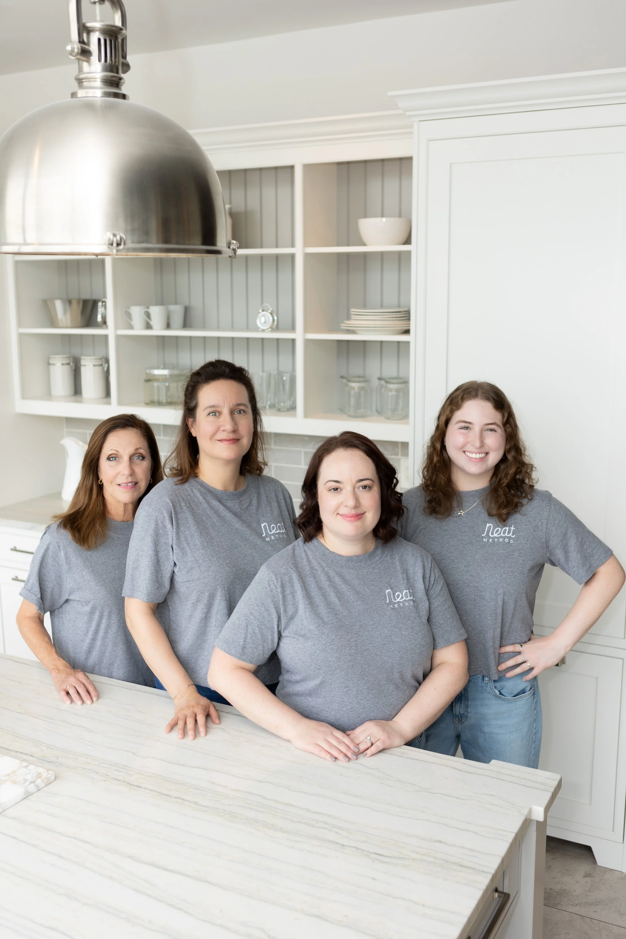 Four women standing in a kitchen, smiling, wearing matching gray shirts with the 'Neat Method' logo, with open white shelves and a marble countertop in the background.
