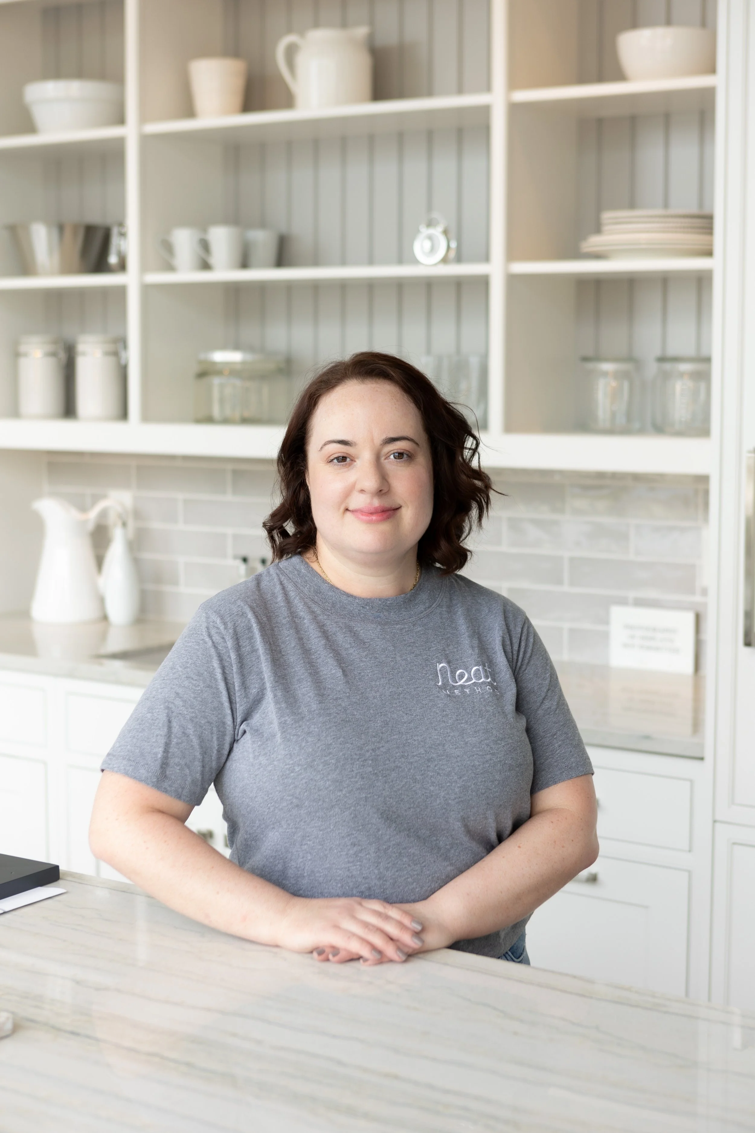 A woman with brown hair and fair skin smiling at the camera in a kitchen. She is wearing a gray t-shirt with a logo on it. The kitchen has open shelves with plates, bowls, mugs, and jars, and a white tiled backsplash.
