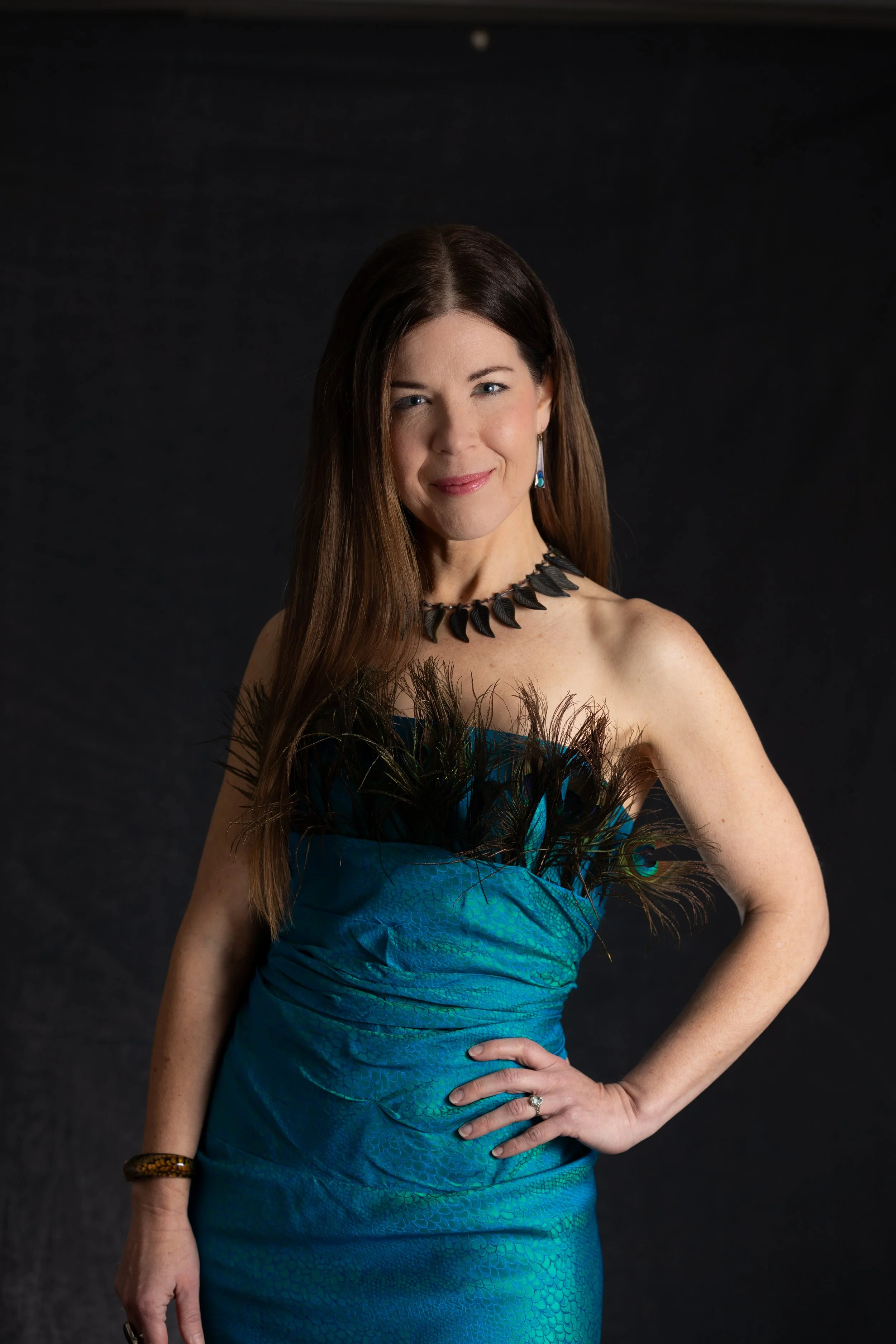 A woman with long brown hair wearing a strapless blue and black dress with feathers, black necklace, turquoise earrings, and a ring on her left hand, posing against a black background.
