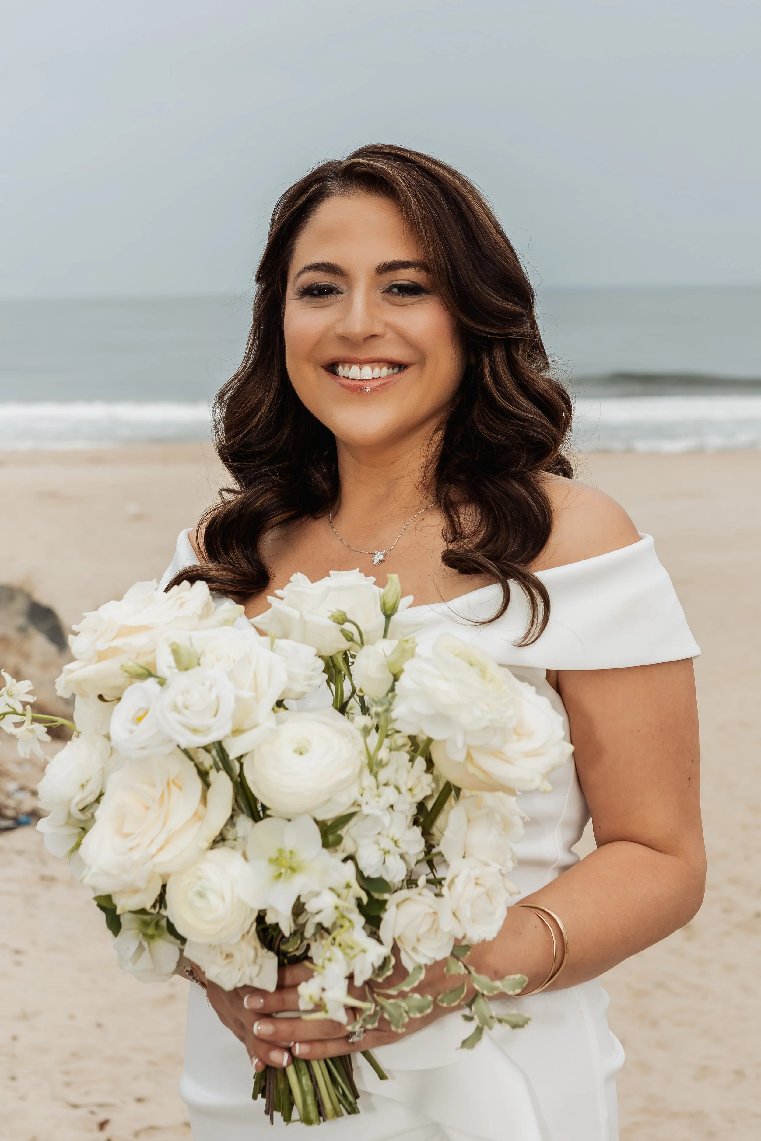 Woman in white wedding dress holding a bouquet of white flowers on the beach.
