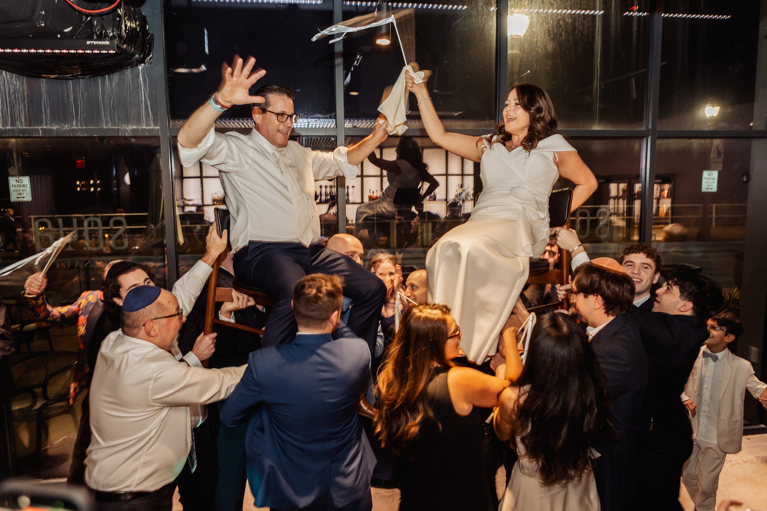 A celebration scene at a restaurant with a woman in a white dress sitting on a chair being lifted by people around her, as a man in white shirt and glasses holds her hand and smiles. The crowd appears joyful, with many smiling and looking at her, and