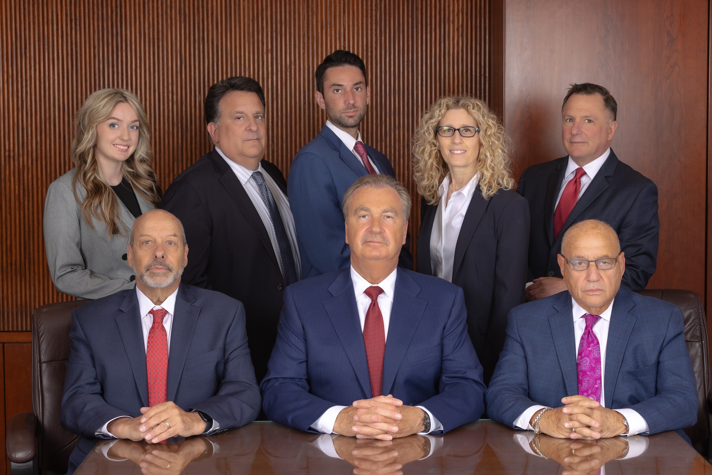 Group of seven individuals, mostly men in suits, seated and standing in a formal setting with a wooden wall background.