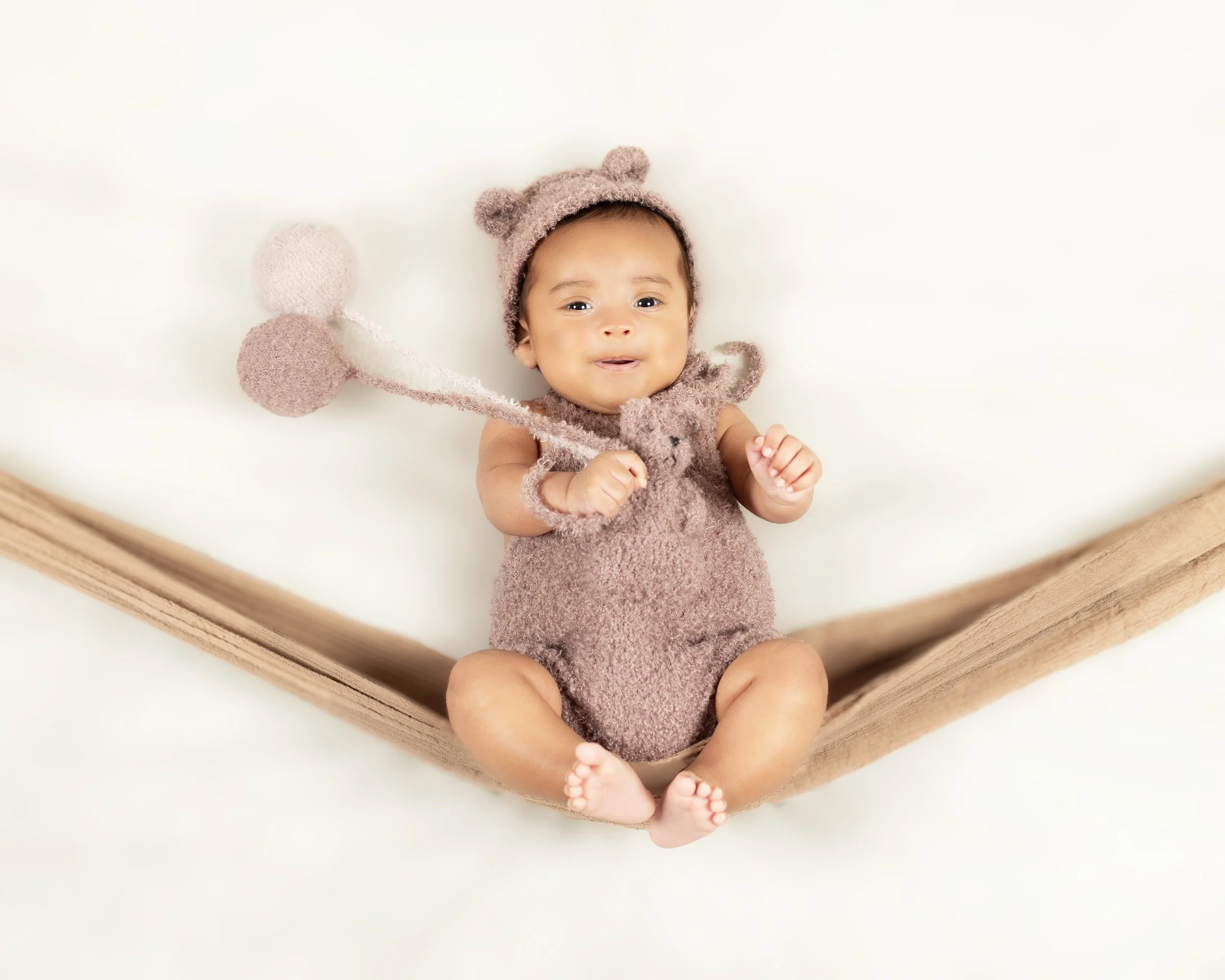 A baby with a brown fuzzy bear costume lying in a wooden crib, holding a pink plush flower toy, with a white background.