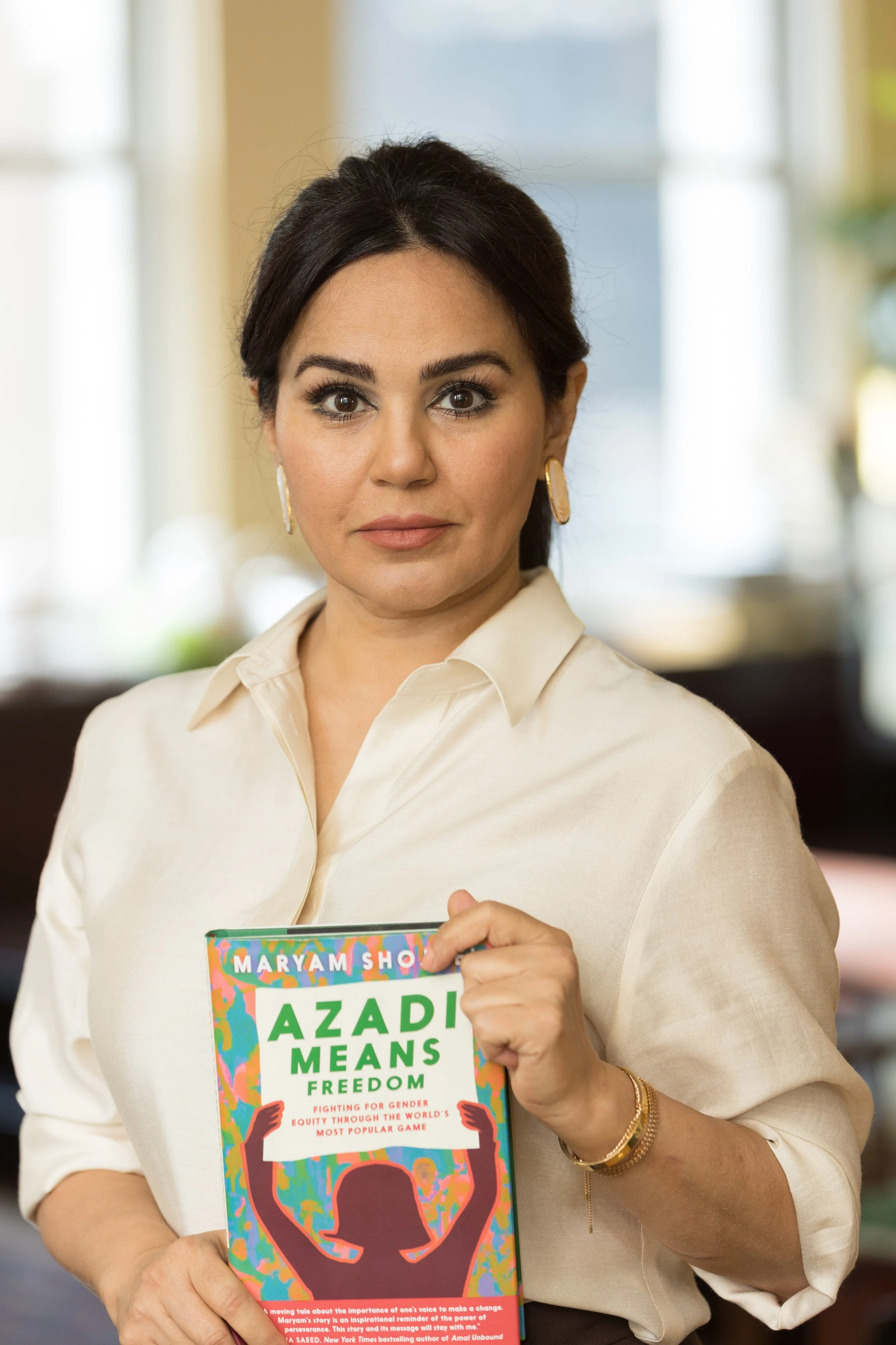 A woman with dark hair, wearing a cream-colored blouse and gold earrings, holding a colorful book titled 'AZADI MEANS FREEDOM' by Maryam  Shojai. She is indoors with blurred windows and greenery in the background.