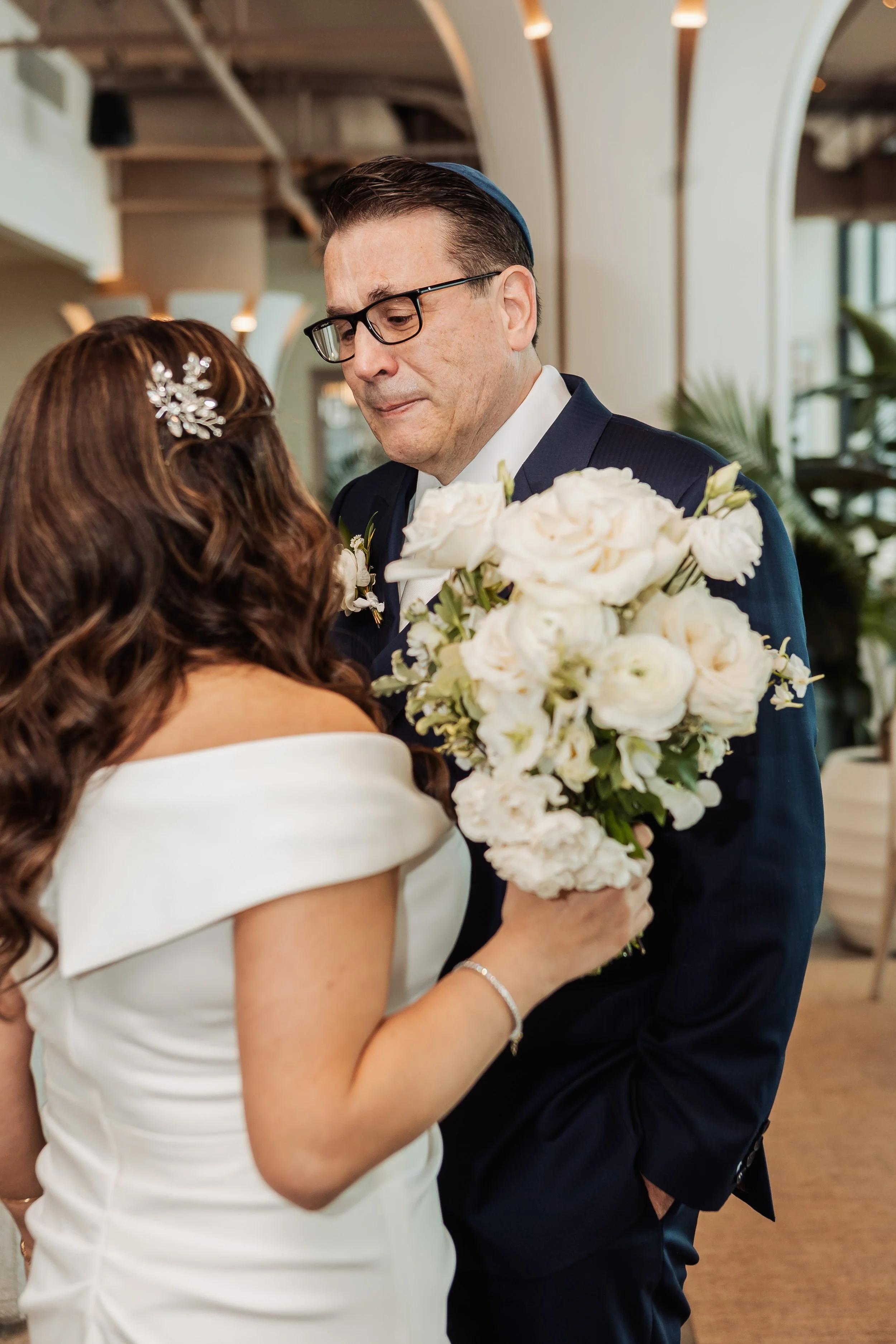 A man in a dark suit and glasses holding a large bouquet of white flowers, looking at a woman in a white dress with a floral hair accessory, in a modern indoor setting.