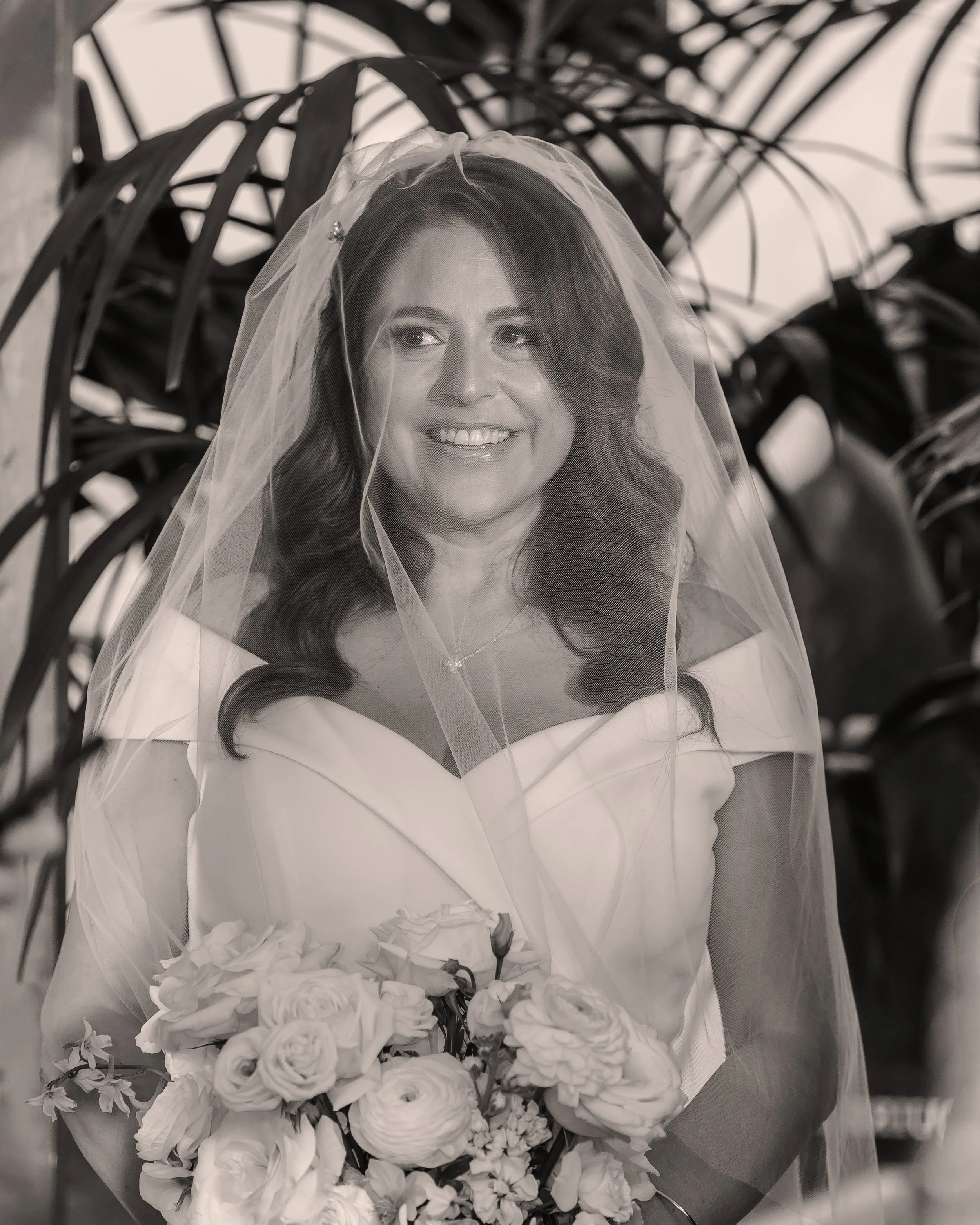 Black and white photo of a woman wearing a wedding veil, smiling, with a bouquet of roses in front of her. The background consists of black metal chairs.