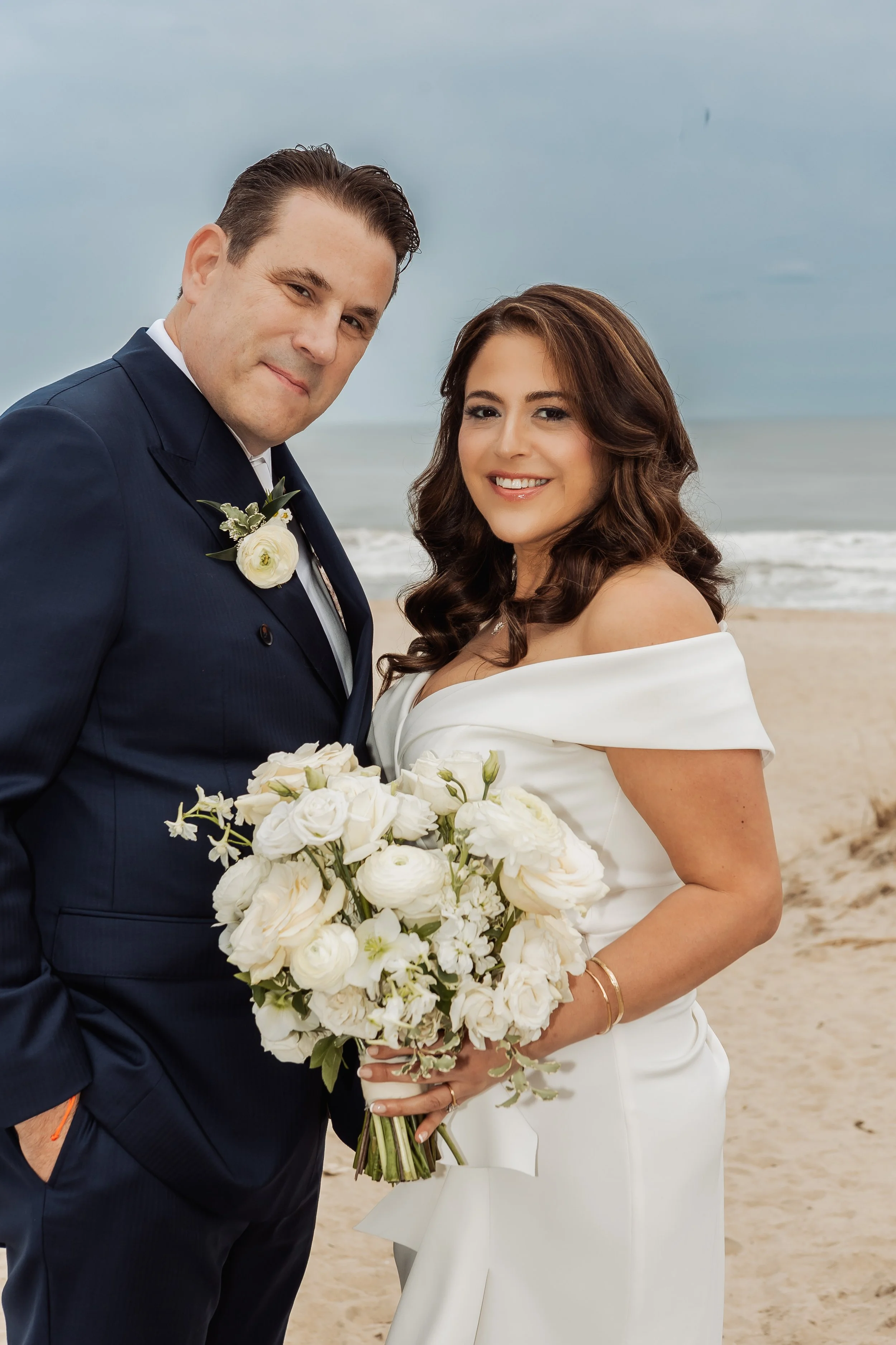 A newlywed couple dressed in formal wedding attire stands on a beach with the ocean in the background. The groom, in a navy suit with a white boutonniere, smiles slightly, while the bride, in a white off-the-shoulder gown, holds a large bouquet of wh