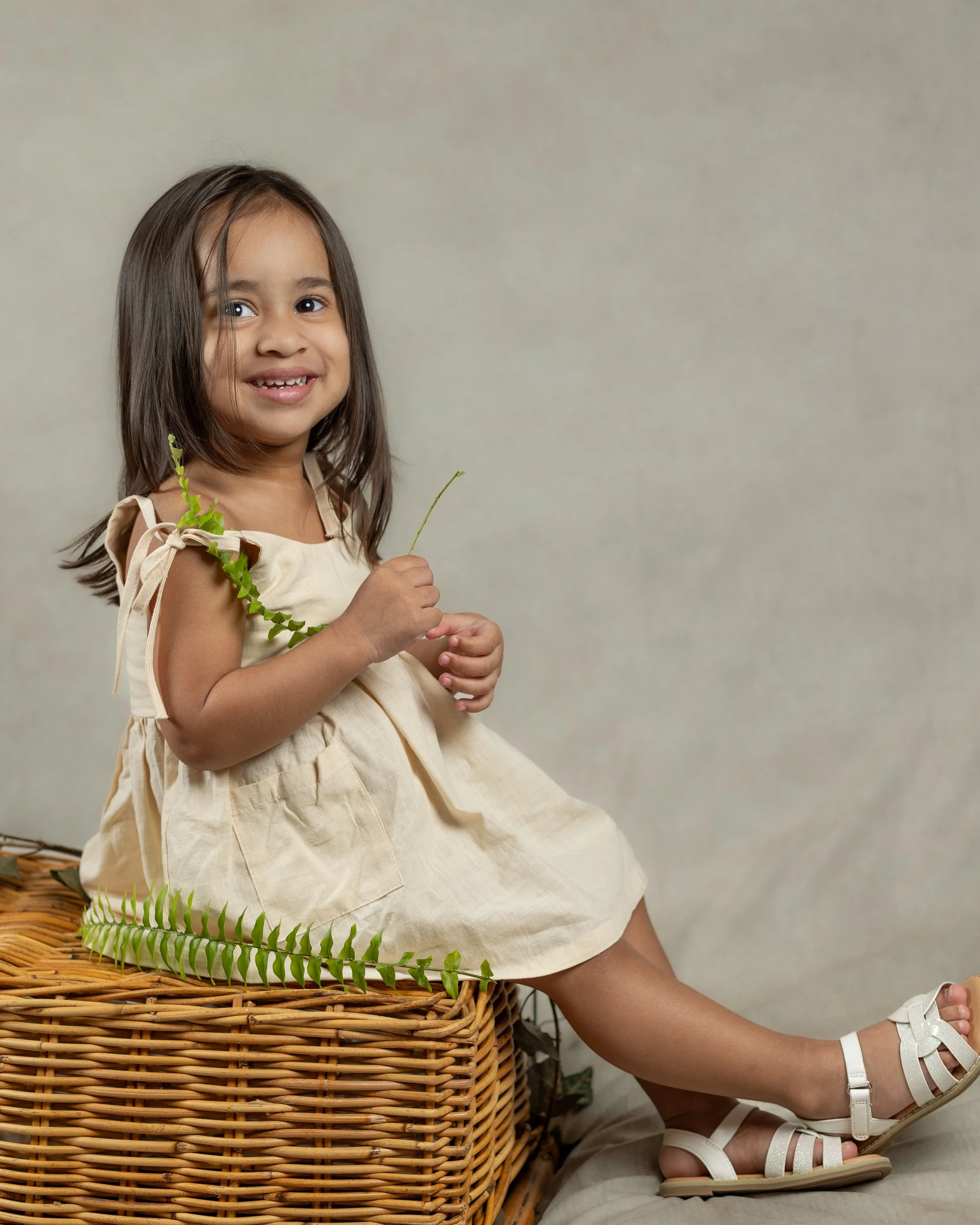 A young girl with long dark hair, smiling, holding a green plant, sitting on a wicker basket with a beige dress, white sandals, and a bracelet, against a plain background.