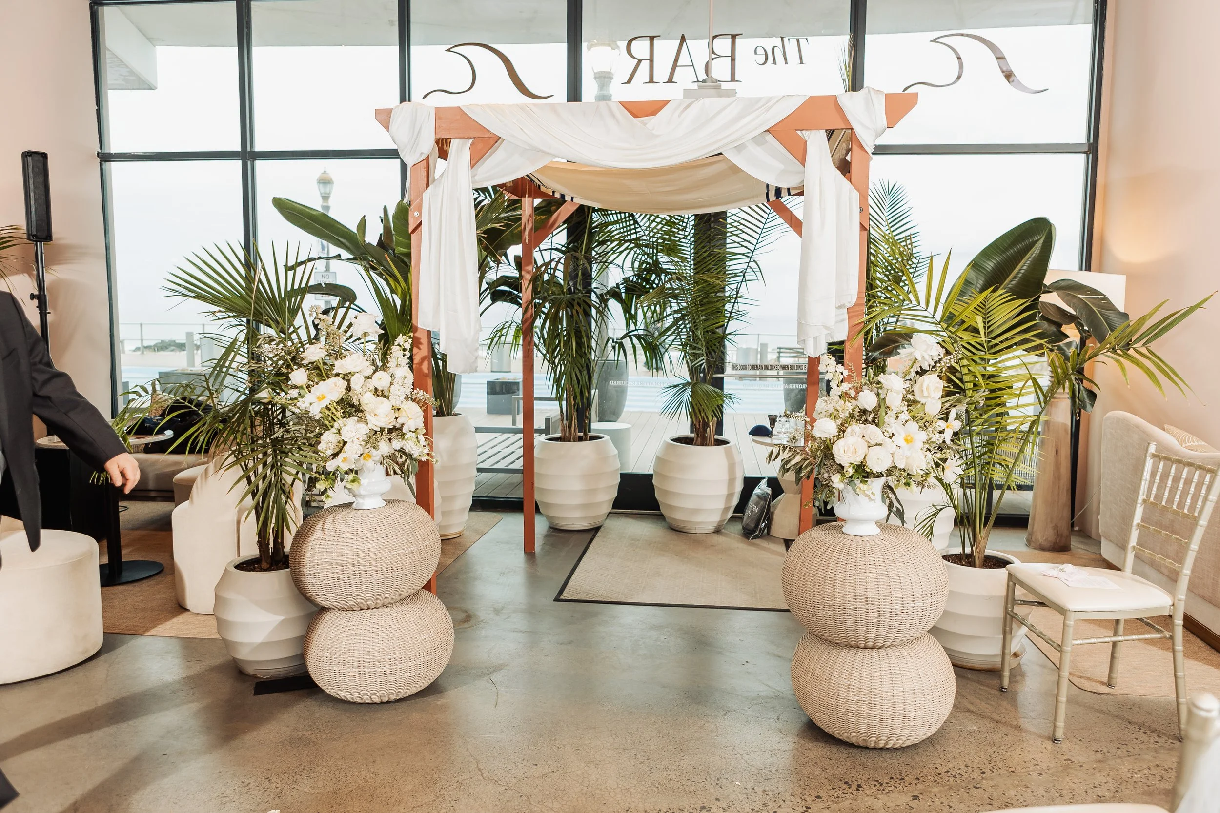 Indoor wedding setup with a wooden arch decorated with white fabric, surrounded by potted plants, white floral arrangements, and wicker stools, with large windows in the background.
