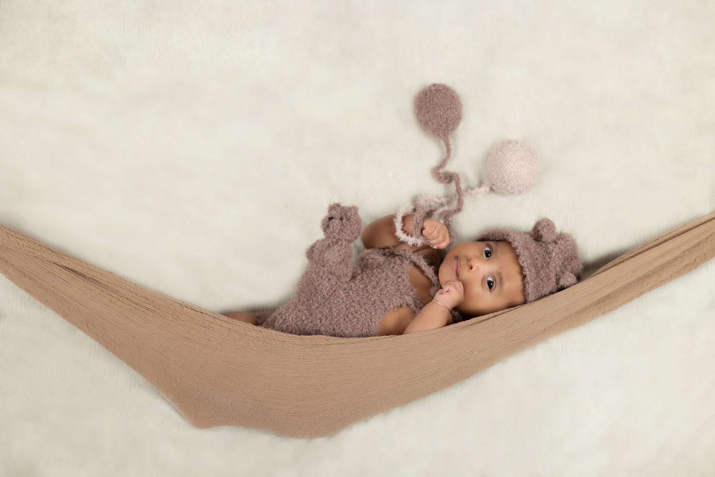 A baby lying in a cozy, beige cradle, dressed in a brown fuzzy outfit with bear ears, holding a light brown teddy bear, on a soft cream-colored background.