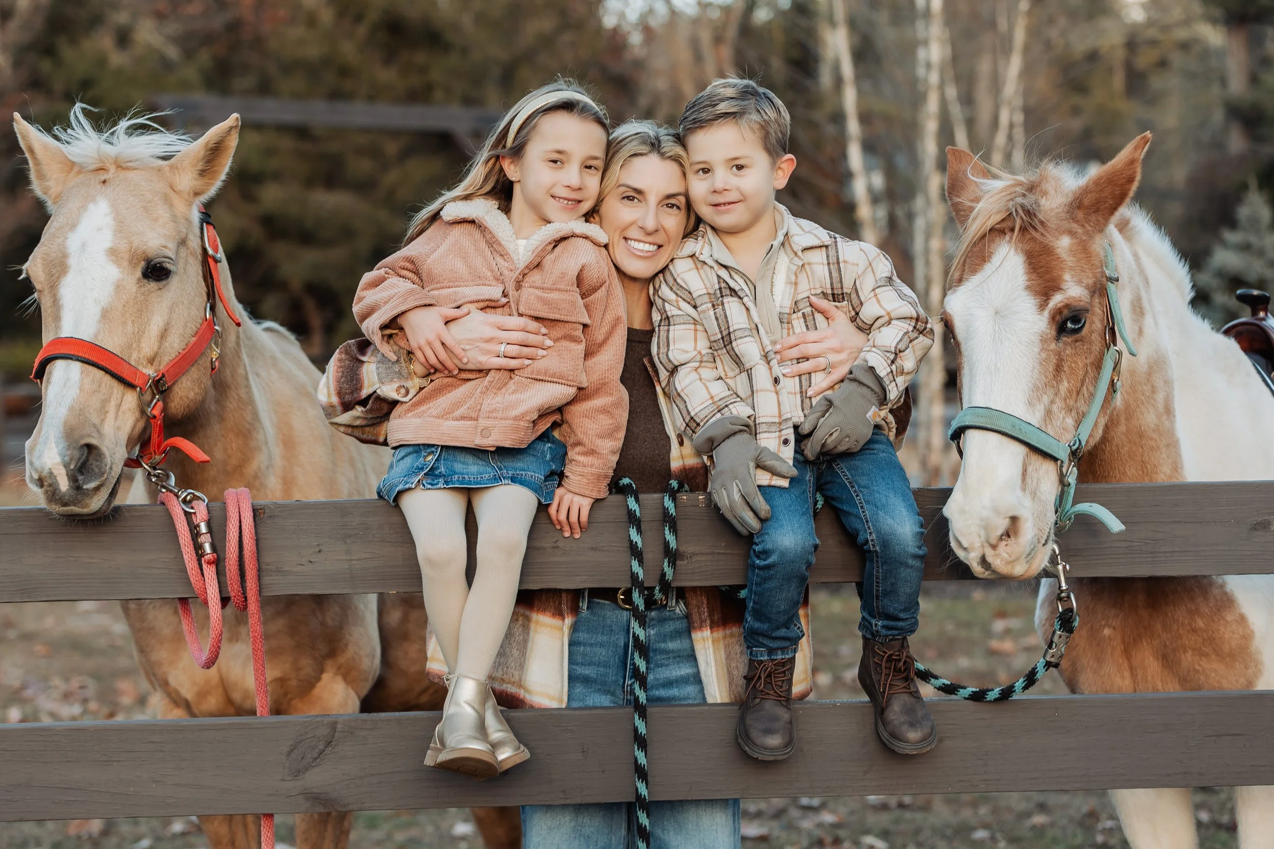 A woman and two children, a girl and a boy, sitting on a wooden fence with two light brown horses, in an outdoor setting with trees in the background, during autumn.