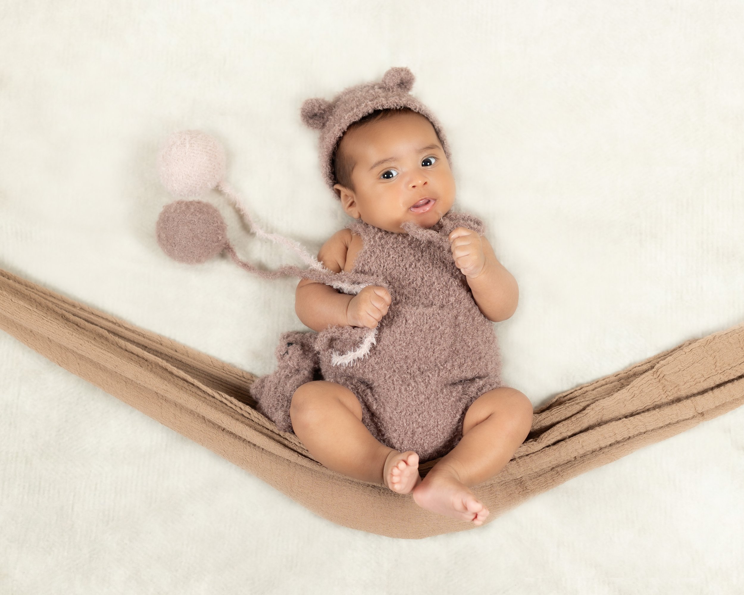 A baby lying on a soft white blanket, wearing a fuzzy brown bear costume with bear ears and a tail, and a brown tail blanket with pink and brown pom-poms attached to a string.