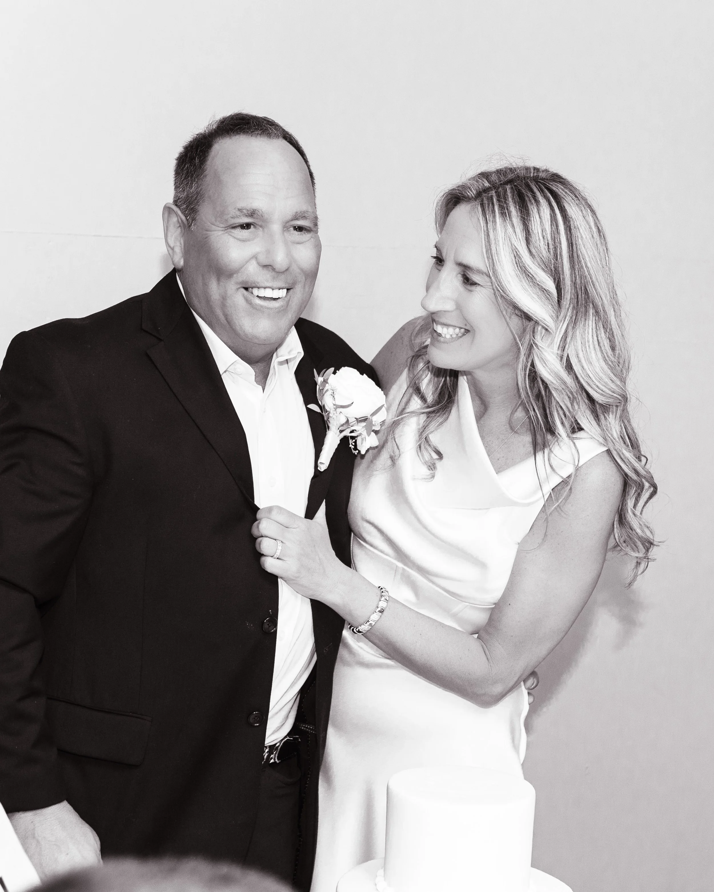 A man in a tuxedo and a woman in a white dress smiling at each other, holding a wedding cake.