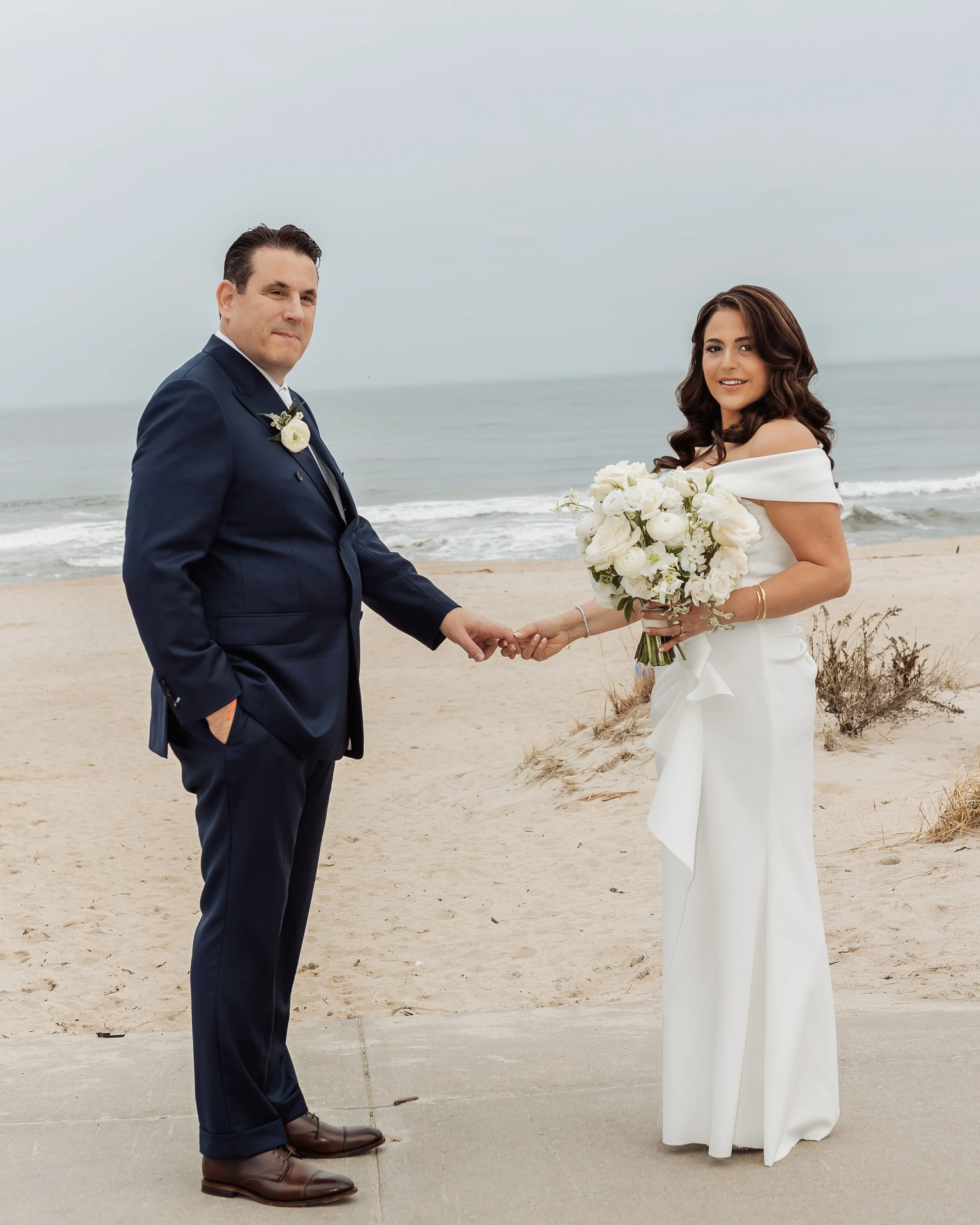 A bride and groom holding hands on a beach, with the ocean in the background. The groom is wearing a navy suit and brown shoes, and the bride is wearing a white off-shoulder wedding dress and holding a bouquet of white roses.
