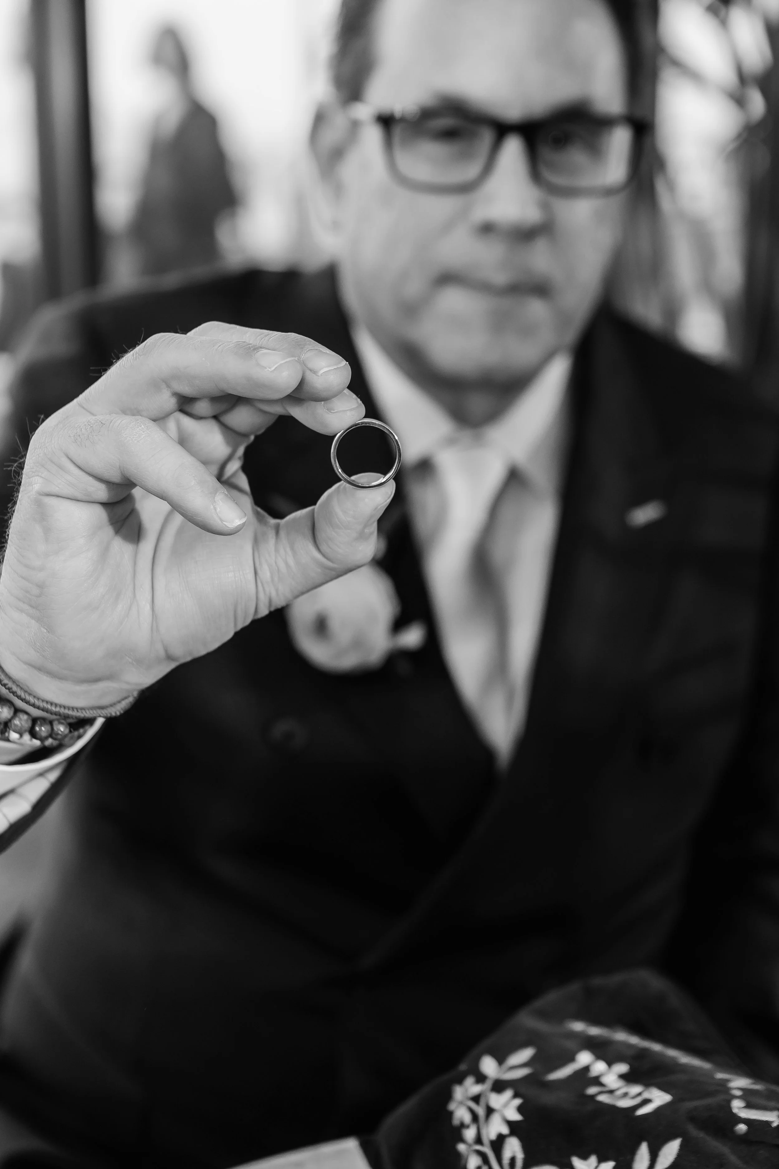 A man wearing glasses in a suit holds a ring towards the camera. The background is blurred, showing a person walking.