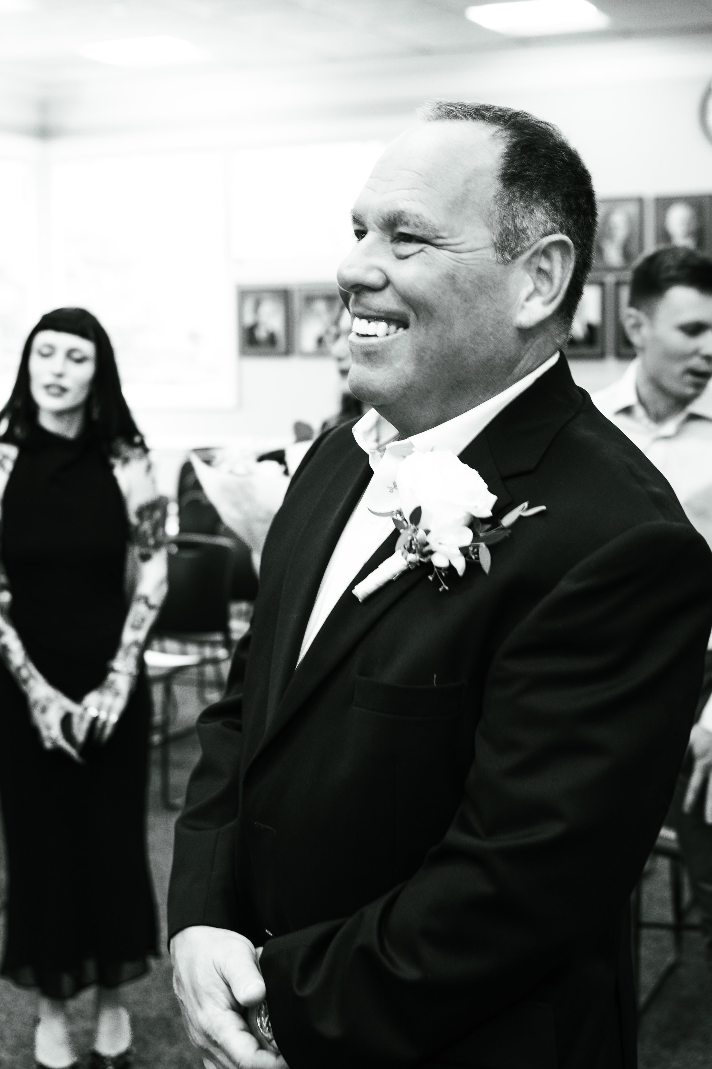 A man in a suit and tie with a boutonniere, smiling and looking to his right, at a social gathering with people and framed pictures on the wall in the background.