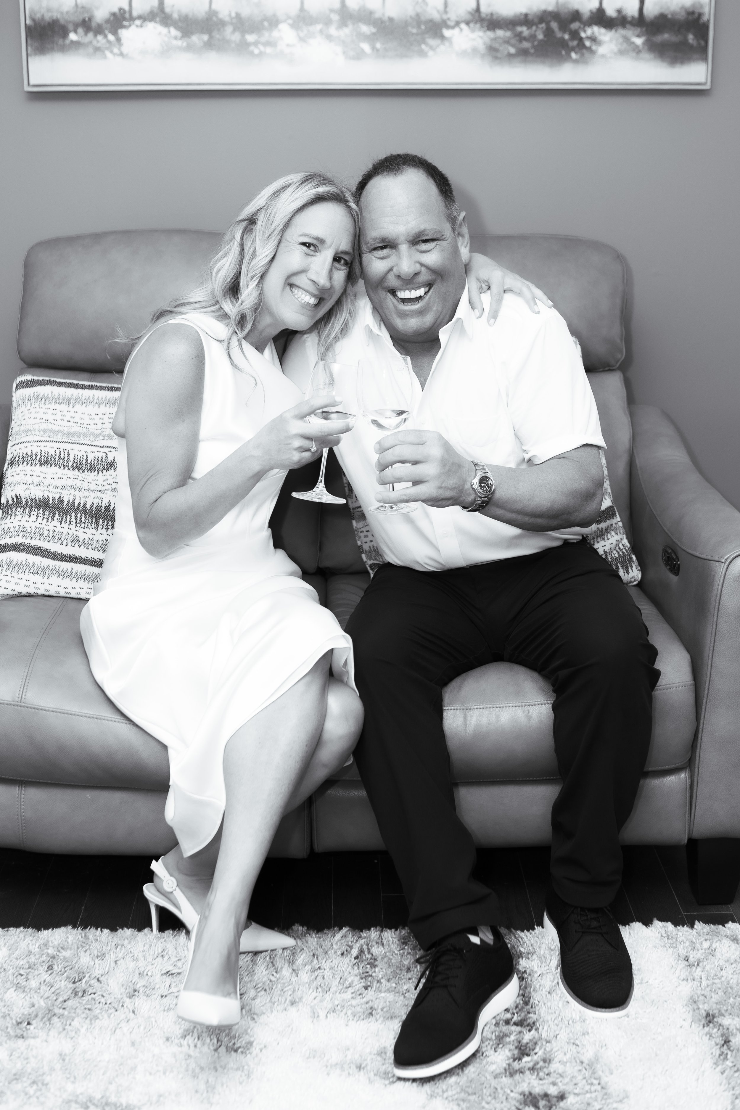 A black and white photo of a smiling couple sitting on a sofa, holding glasses of wine, with the woman's arm around the man's shoulder, in a cozy living room setting with a picture frame on the wall behind them.