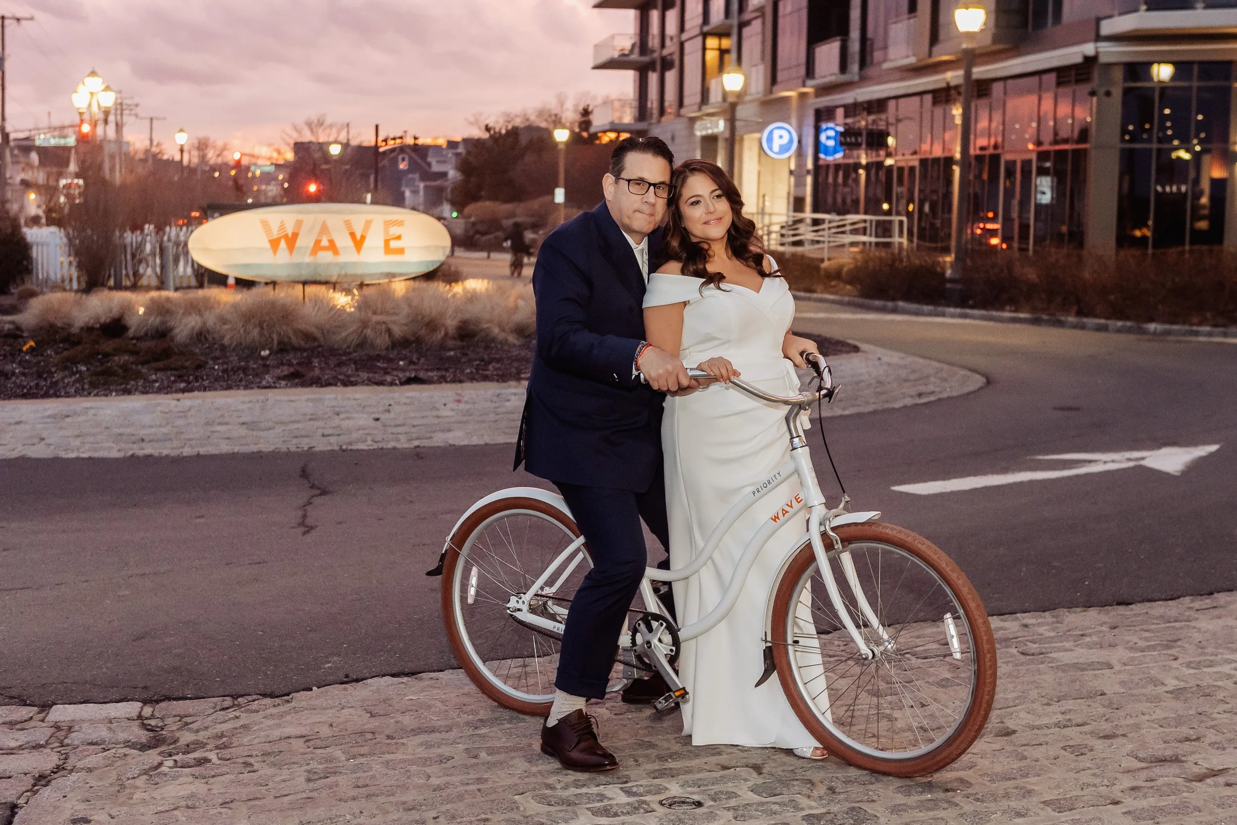 A woman in a white off-the-shoulder dress riding a white bicycle with a man in a black suit sitting on the handlebars during sunset in an urban area with modern buildings, street lamps, and a sign reading 'WAVE'