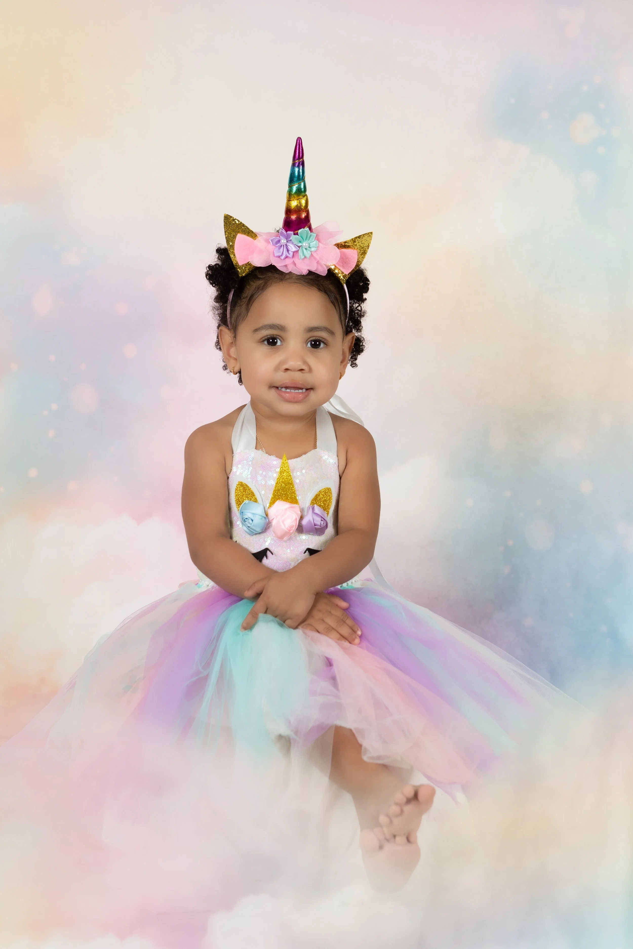 A young girl sitting on a cloud-colored background, wearing a rainbow unicorn headband and a colorful tutu dress, smiling softly.